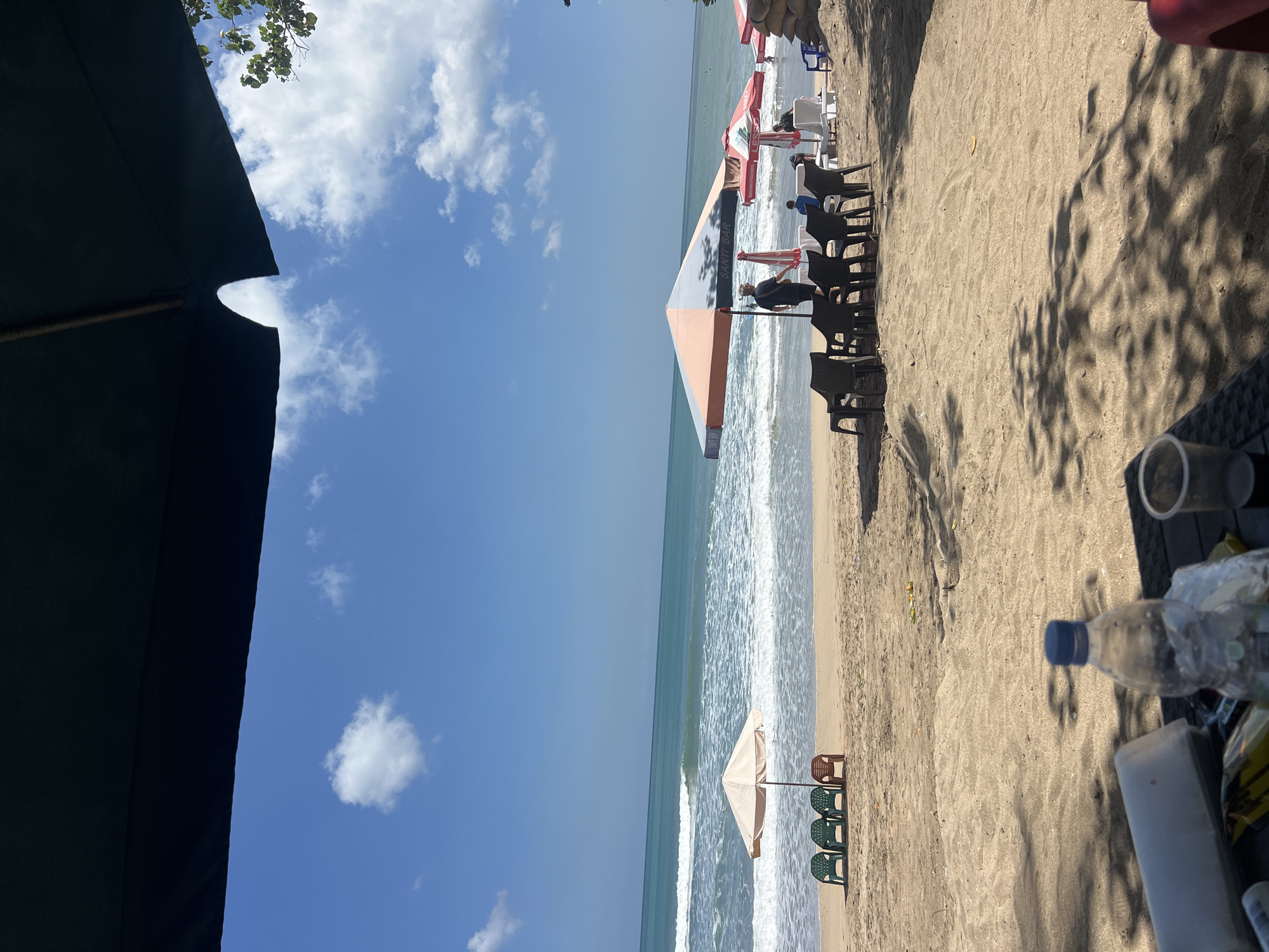 This is an eye-level, medium shot of Kuta Beach in Kuta, Indonesia, on a sunny day with a clear blue sky and scattered white clouds. The foreground is dominated by sand with dappled shadows from palm trees. On a dark-colored table in the immediate foreground, a plastic bottle of water, a white paper cup, and a pack of cigarettes are visible. The middle ground features a sandy beach with rows of chairs and beach umbrellas. To the left, a small, light-colored umbrella is set up with four plastic chairs in front of it: three green and one red. Further to the right, a larger, two-toned umbrella (orange and gray) shades a group of black plastic chairs and a table. Behind this umbrella, several red and white striped beach umbrellas are visible, along with lounge chairs and people. A man in a dark t-shirt and shorts stands near the large umbrella. In the background, the turquoise ocean meets the sky, with gentle waves rolling onto the shore. Several people are visible in the water, some appearing to be surfing. The overall impression is a relaxed beach scene.