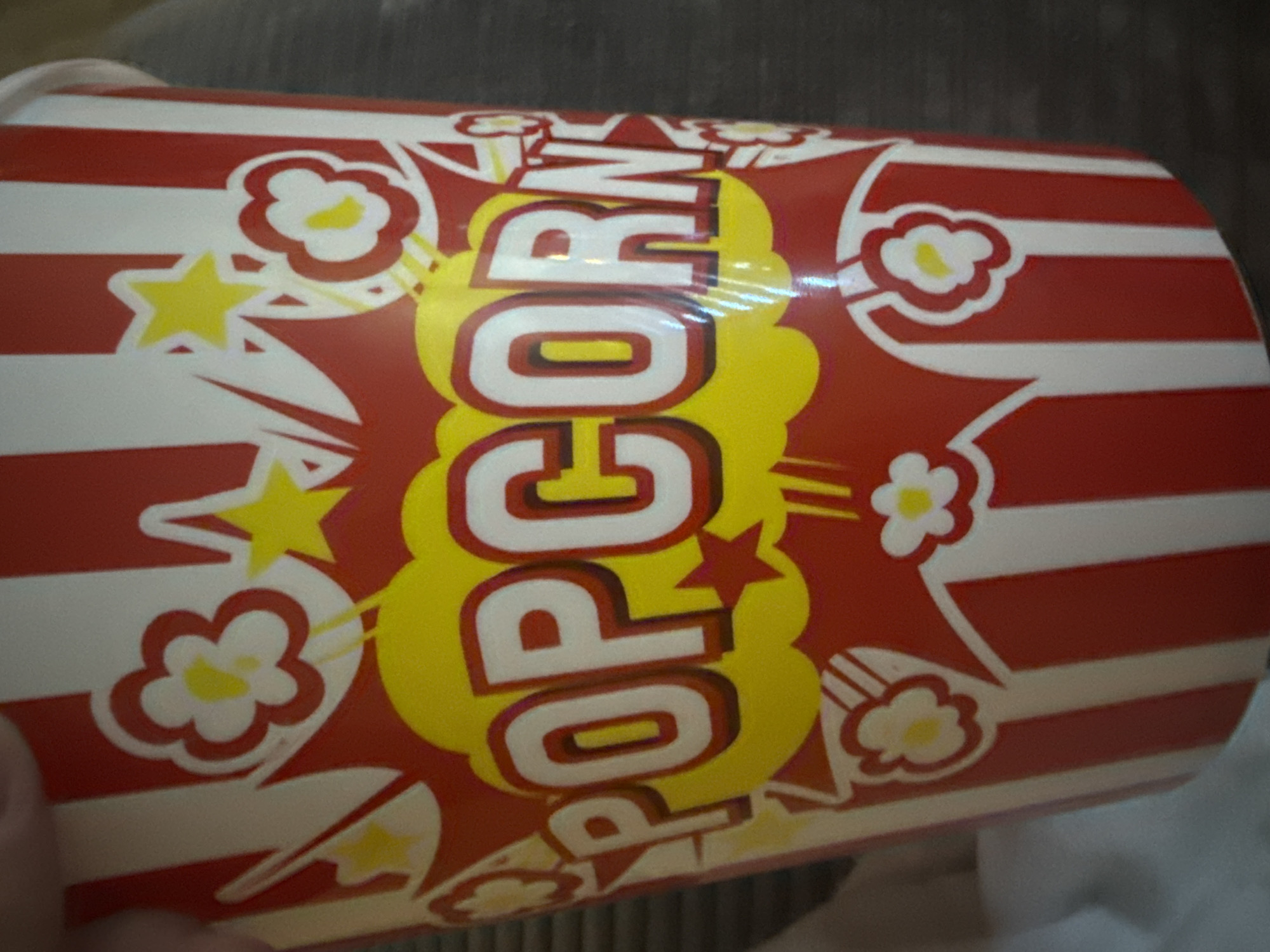 The image shows a close-up of a red and white striped popcorn bucket with yellow accents and cartoonish white flowers and stars. The word "POPCORN" is prominently displayed in large, white, block letters with a red outline on a yellow burst shape. The bucket is cylindrical and appears to be made of paper. The background is a blurred gray surface, possibly a table or a piece of furniture. There is no indication of the location in the city of Varna, Bulgaria.