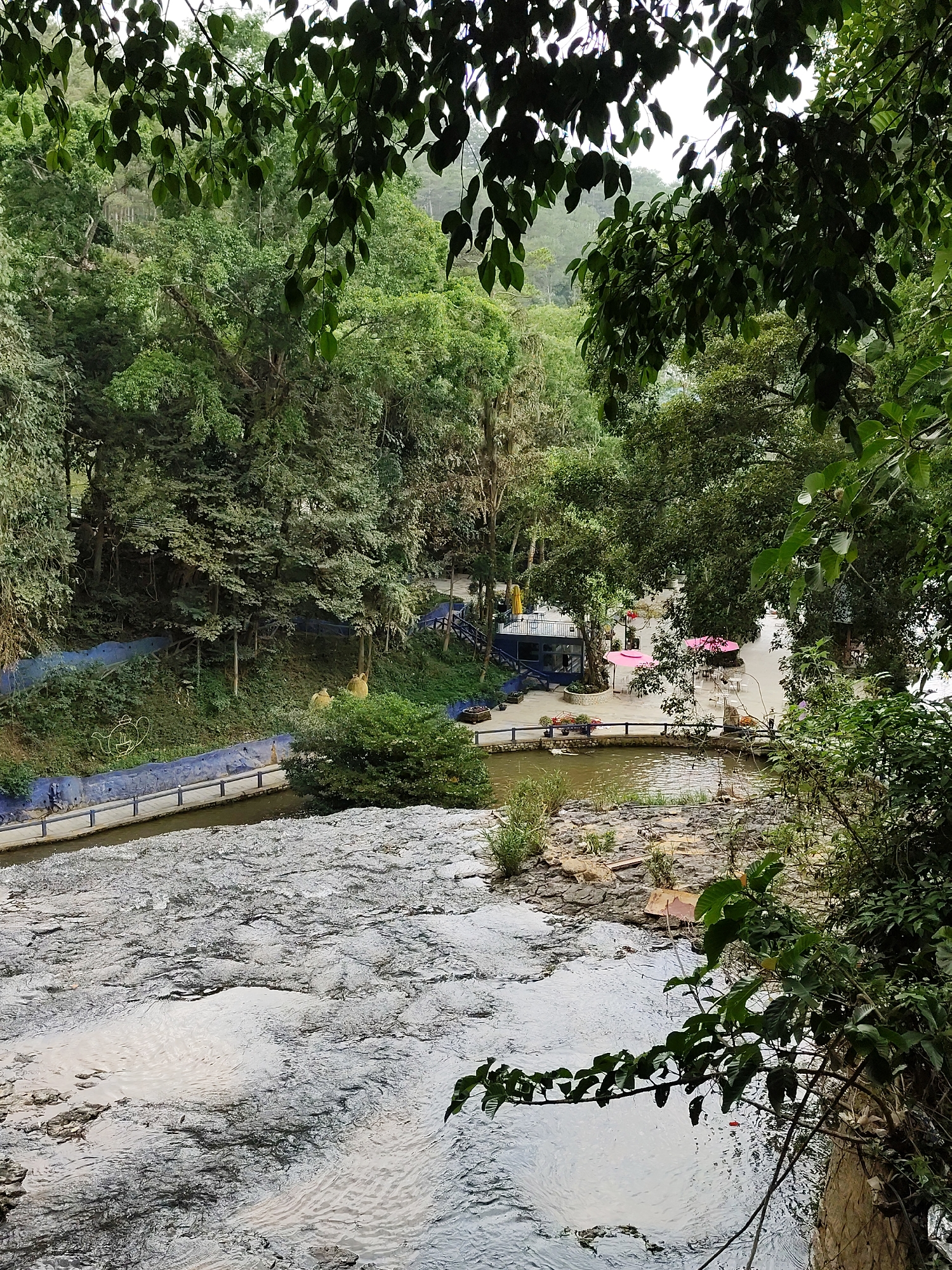 The photograph presents a view of Datanla Falls in Da Lat, Vietnam, seen from an elevated vantage point through lush vegetation. The cascading water flows over layered rock formations. A developed area with a blue structure, tables with pink umbrellas, and walkways is visible to the right of the waterfall. People are present in the developed area. A blue wall adorned with decorations and a bridge runs along one side of the waterway above the falls. Foliage frames the scene.