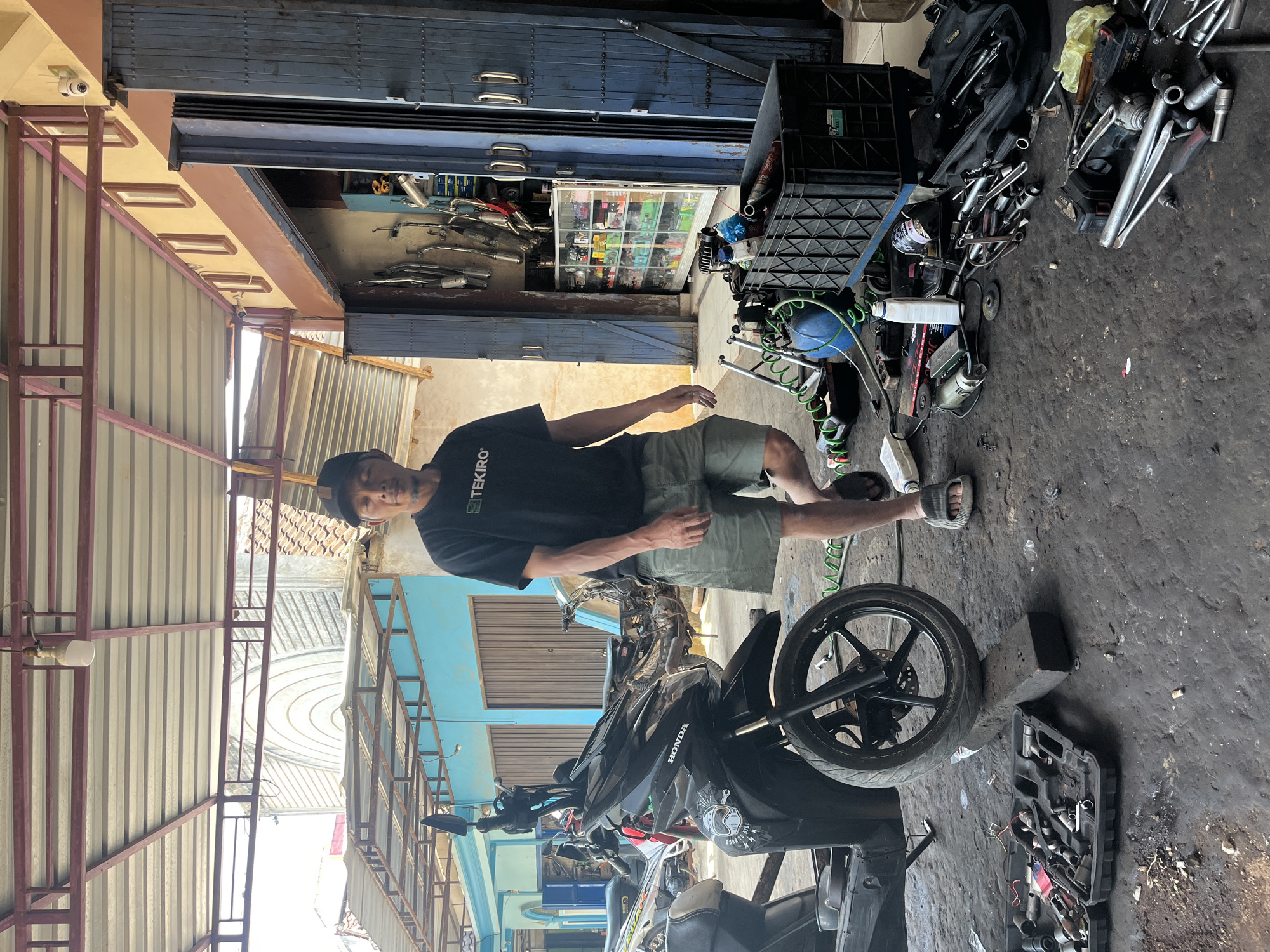 A man stands in a motorcycle repair shop in Karang Sari, Indonesia. He is wearing a black t-shirt with "TEKIRO" printed on it, olive green shorts, a black baseball cap, and grey sandals. He is standing next to a black Honda scooter, which is parked on a wooden block. The shop appears to be open, with tools and parts scattered on the concrete floor. In the background, there is a shelf filled with motorcycle parts and a blue folding door. The scene is illuminated by natural light.