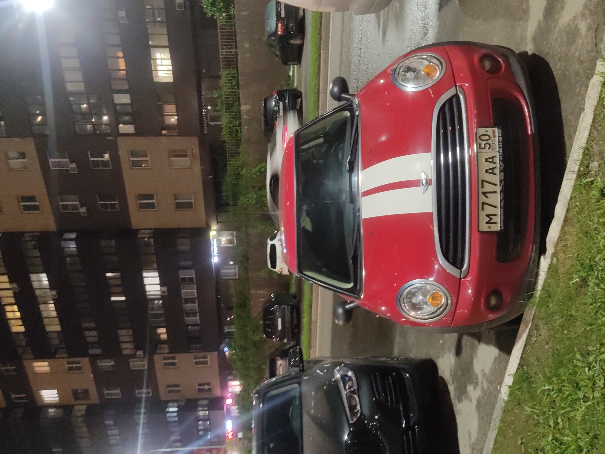 The image depicts a street scene at night in an urban area of Unknown, Unknown. A tall building with numerous windows dominates the left side of the frame, illuminated by streetlights. Several cars are parked along the right side of the street. A red Mini Cooper with white racing stripes is prominently positioned in the foreground, partially parked on the sidewalk. Its license plate, "M 717 AA 50 RUS," is visible. Adjacent to the Mini Cooper is a black vehicle, presumably a car, with its headlights visible. The street appears wet, and there is vegetation (grass) along the curb.