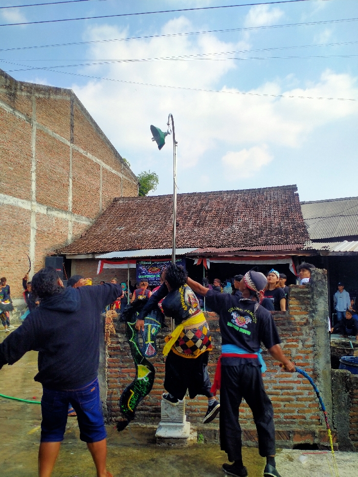 The image captures an outdoor cultural performance in Sepat, Indonesia, under a blue sky with scattered clouds. In the foreground, three males are actively engaged. The central figure, dressed in a black top with a yellow patterned sash and black pants, is in mid-air, seemingly interacting with a tall metal pole topped with a green, inverted-cone fixture. This individual is connected to a large, dark green, serpentine-like object with geometric patterns. To the left, a male in a black hoodie and blue shorts stands with arms outstretched. To the right, another male in a black t-shirt, black pants, and a blue and red sash holds a multi-colored braided whip.
Behind the performers, spectators observe from various positions, including behind a low brick wall and further back near the buildings. The background features a large, unfinished brick building on the left, characterized by exposed concrete beams. To the right, a traditional house with a reddish-brown tiled roof is visible, beneath which hangs a red and white banner, indicative of the Indonesian flag. Several overhead power lines traverse the upper portion of the frame. The ground beneath the performers appears to be a paved or concrete surface, exhibiting some wetness in the foreground.