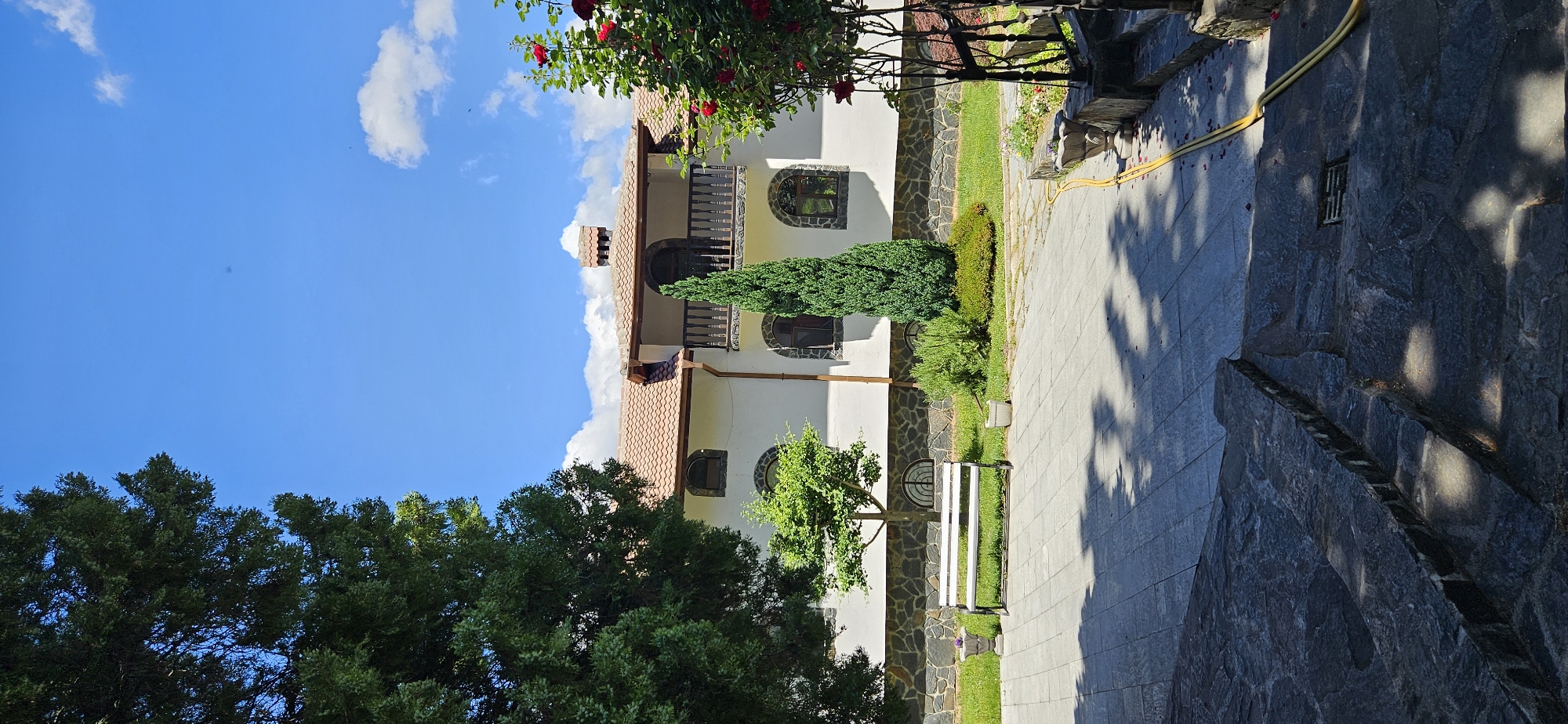 The image depicts a two-story building with a tiled roof, reminiscent of traditional architecture, set against a bright blue sky with scattered clouds. The building has white walls, dark-trimmed arched windows, and a balcony on the upper floor. In front of the building, there is a well-maintained garden with a white bench, a small tree, a tall, slender evergreen, and various shrubs. The foreground features a paved walkway made of large, grey stones, with dappled sunlight and shadows cast by overhead trees. To the right of the walkway, a yellow hose is visible, snaking along a stone wall. The overall scene suggests a peaceful, possibly rural or suburban setting, and the architecture hints at a location in Bulgaria, potentially the village of Ovchartsi, given the context. There are no people or animals visible in the image.