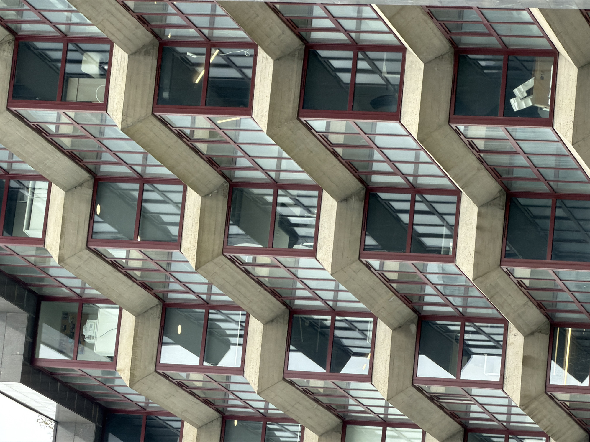 The image shows a close-up of a modern building's facade, characterized by a grid of windows set into a concrete structure. The windows have red frames and multiple panes, reflecting the sky and some interior elements. The concrete is light gray and textured. Visible inside some windows are office interiors, including furniture, lamps, and other items. The image does not contain any people. The building style suggests brutalist architecture. The location, as mentioned in the prompt, is Porto, Portugal.