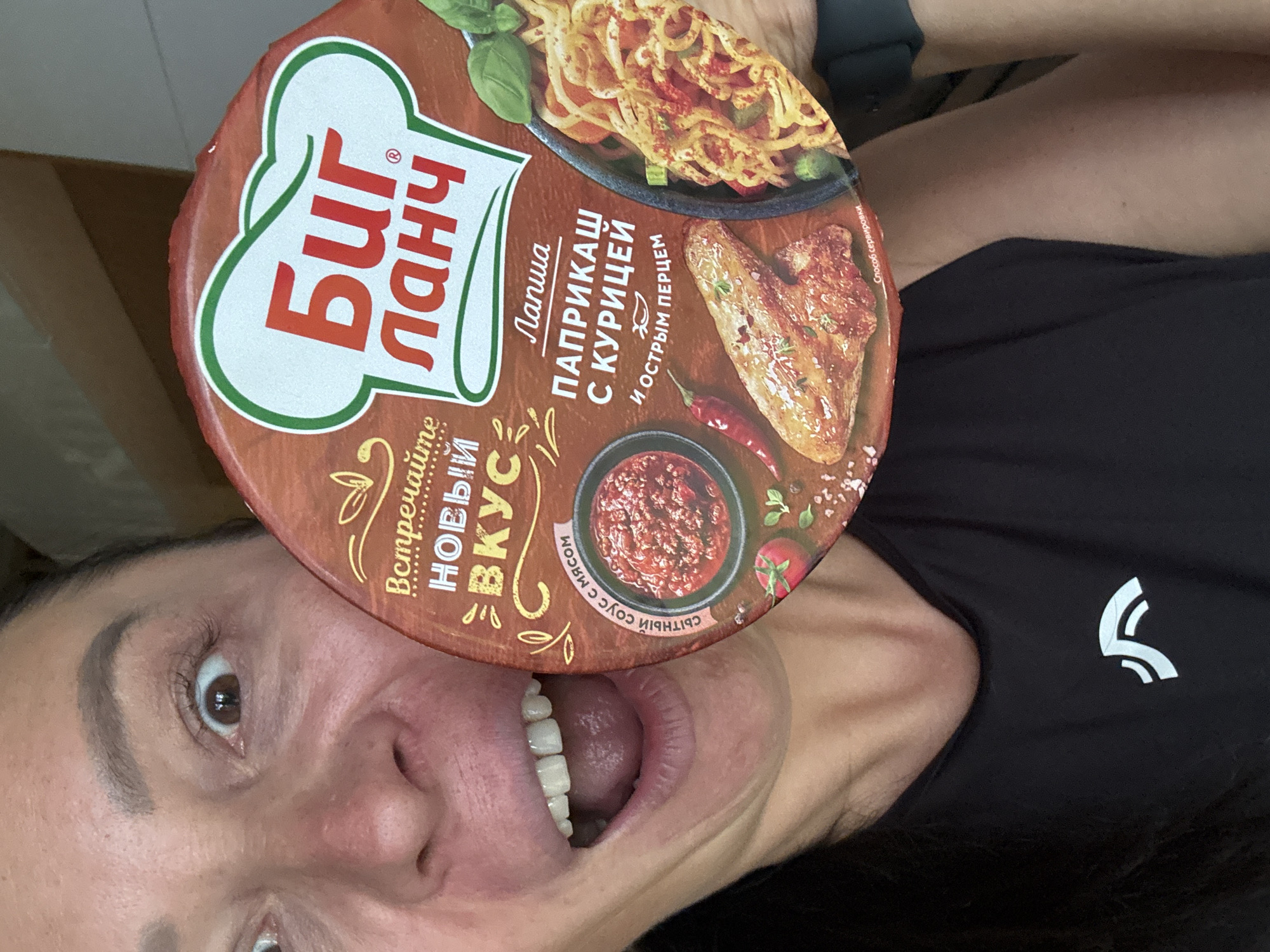 A woman is pictured holding a container of "Big Lunch" instant noodles in front of her face with her mouth open. The container shows images of noodles with chicken and a spicy sauce. The text on the container is in Russian. She is wearing a black shirt with a white logo. The photograph appears to be taken in an indoor environment. The city is Chaikovsky, Russia.