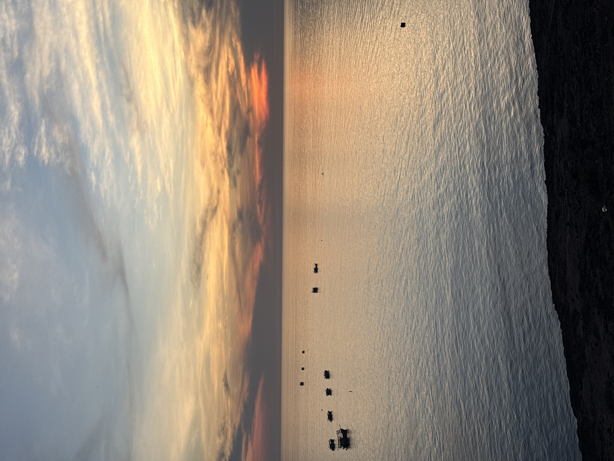 A sunset over the ocean near Lý Sơn Island, Vietnam, casts a warm, golden glow across the water, with scattered boats dotting the calm surface. The sky is a canvas of soft blues, oranges, and pinks, with wispy clouds painted across the horizon. In the foreground, the dark, rocky coastline of the island provides a stark contrast to the serene seascape.