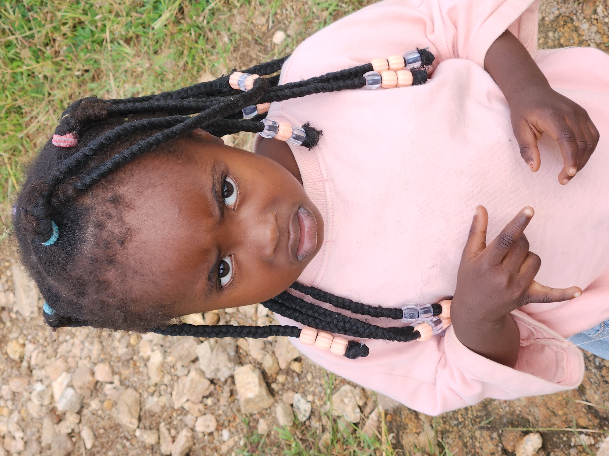 A young Black girl with dark skin and dark hair braided with blue and pink beads is looking directly at the camera with a stern expression. She is wearing a light pink long-sleeved sweatshirt and her hands are in front of her chest, one with fingers extended. The background consists of loose rocks and green grass.