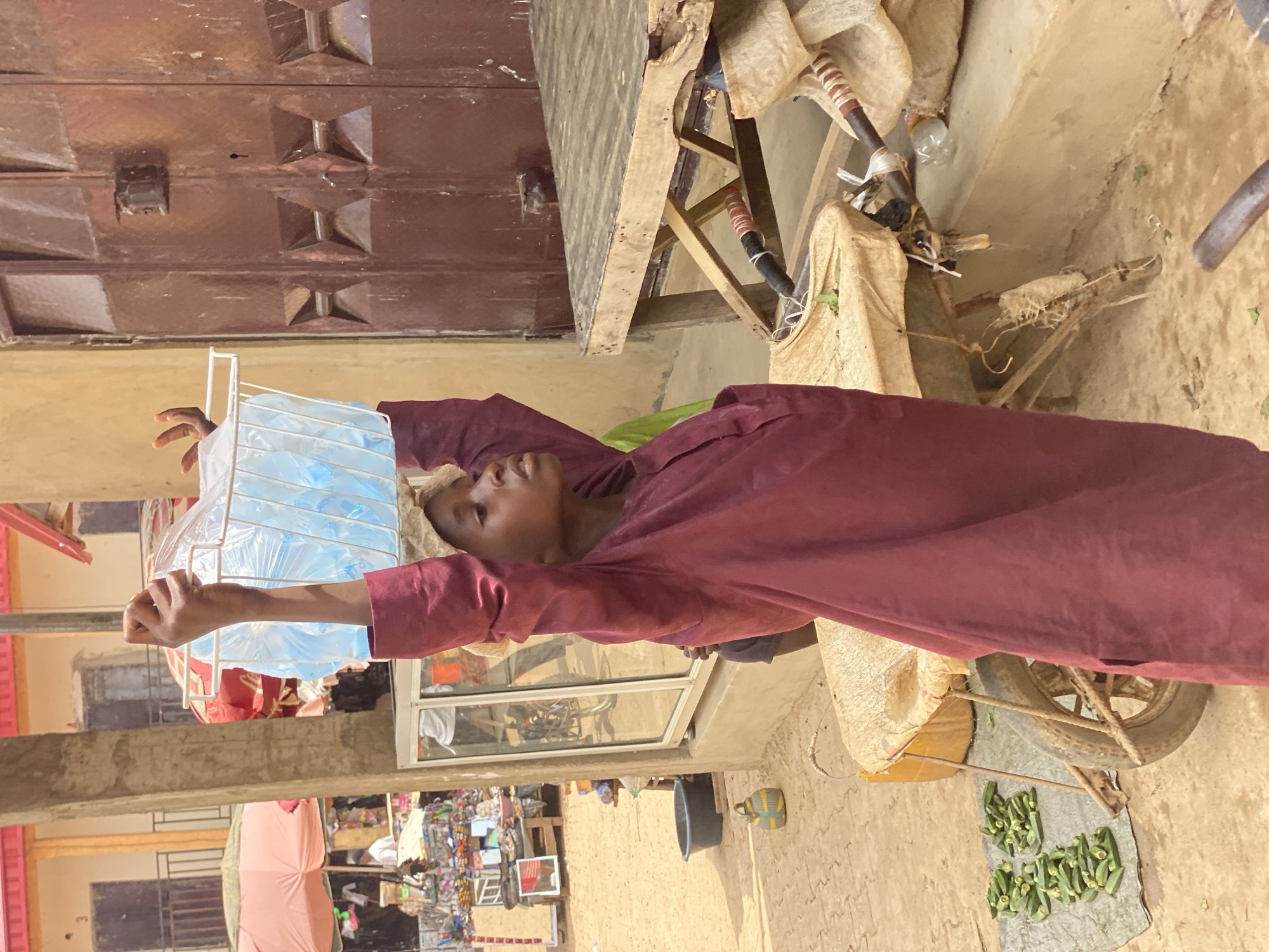In Kafanchan B, Nigeria, a person in a maroon robe is seen holding a white wire rack filled with blue water pouches. The individual stands in front of a storefront with glass windows, beyond which a bustling market scene is visible. A bicycle modified with a wooden platform is beside the person. The platform is covered with a beige cloth. Okra vegetables are arranged on a grey cloth near the bicycle's rear tire. Above, a brown door with rectangular designs is situated.