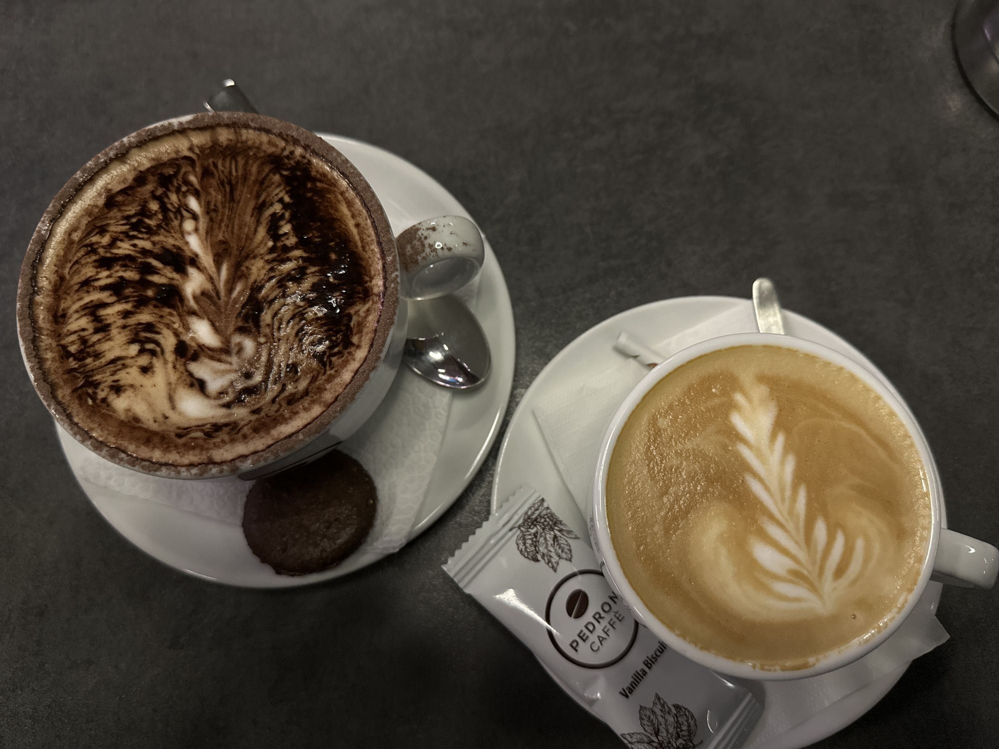 Two cups of coffee are on a dark grey surface. The cup on the left contains a cappuccino with dark brown and white foam, presented with a small round chocolate biscuit and a silver spoon on a white saucer over a white napkin. The cup on the right holds a latte with a latte art design of a fern. It also sits on a white saucer with a white napkin, and a silver spoon. Next to this cup is a white packet labeled "PEDRON CAFFE" and "Vanilla Biscuit". The scene appears to be in a coffee shop or cafe. The location, Stara Zagora, Bulgaria, is not discernible from the image itself but may be inferred from the coffee brand, Pedron Caffe, which has a presence in Bulgaria.