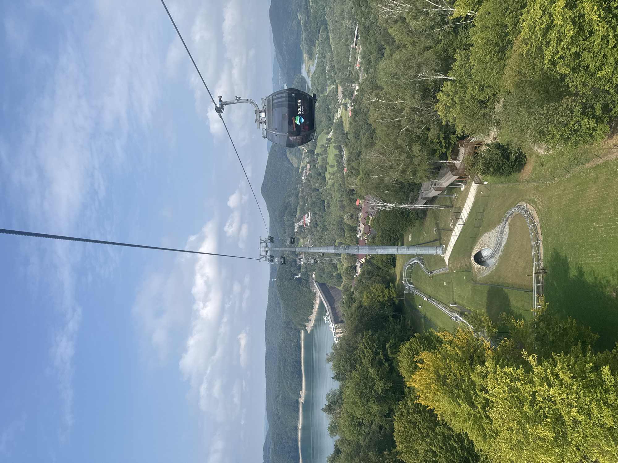 This is a vertical, eye-level, medium shot of a cable car suspended from a steel cable, moving across a lush, green, mountainous landscape. The cable car, identified by the word "SOLINA" printed on its exterior, carries several passengers. In the background, a large, curved dam with a concrete structure is visible across a calm body of water. The dam is surrounded by densely forested hills that fade into the hazy distance. In the foreground, a metal roller coaster track winds through a grassy area, with a tunnel visible at its base. A wooden walkway, elevated and winding, is also present amidst the trees. The sky is a vibrant blue, dotted with fluffy white clouds, suggesting a clear, sunny day. The overall scene depicts a recreational area in Solina, Poland, likely a tourist attraction that combines scenic views with adventurous activities.