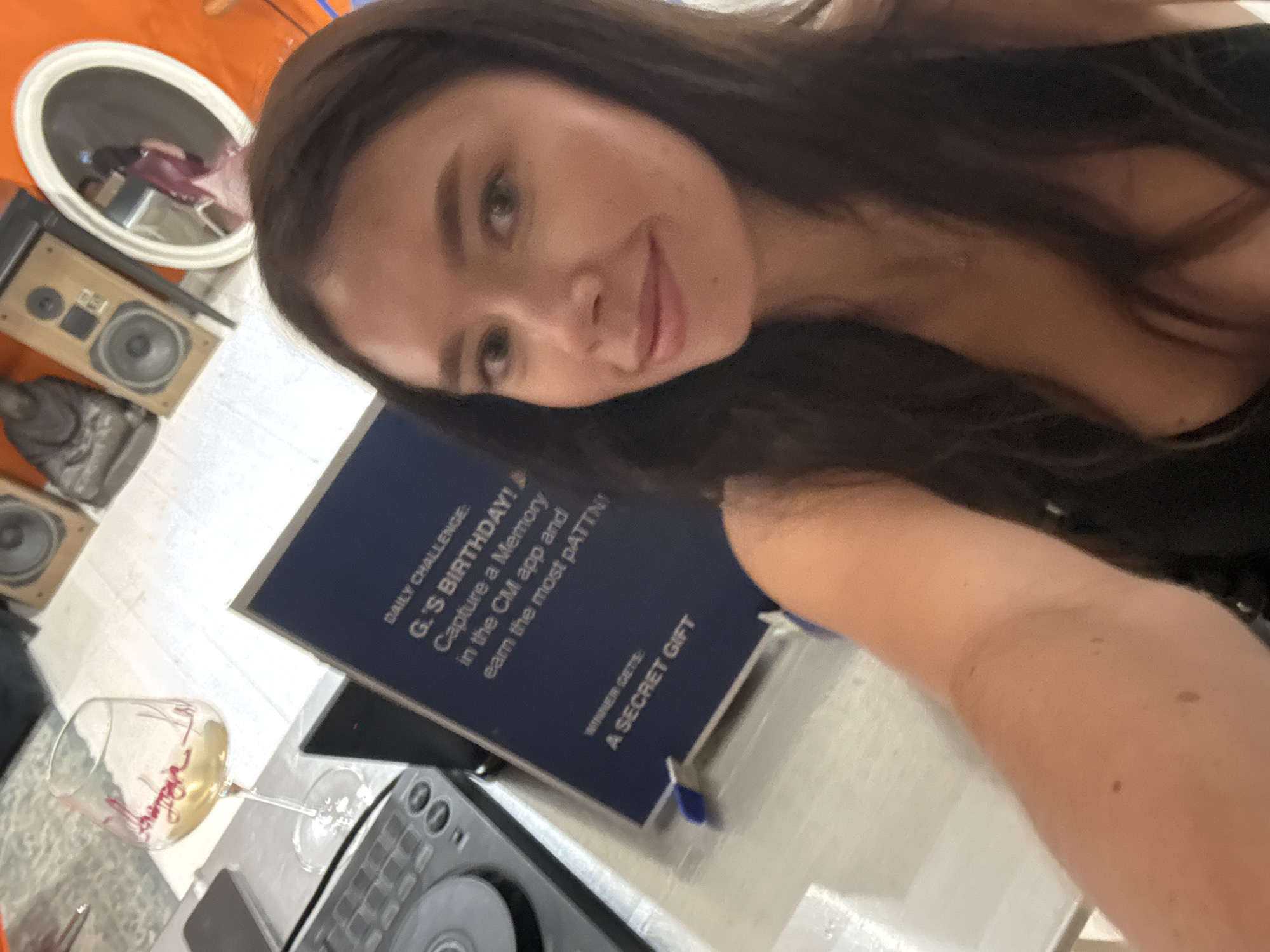 A woman with long, dark hair and light skin is taking a selfie in what appears to be a casual indoor setting. She is wearing a dark top. In the background, to her left, is a wine glass with a light-colored liquid and pink lettering, a DJ turntable, and a dark blue sign with white text that reads "DAILY CHALLENGE: G.'S BIRTHDAY! Capture a Memory in the CM app and earn the most paTTN! WINNER GETS: A SECRET GIFT." To her right, in the background, are two speakers, a statue of a Buddha, and a round mirror reflecting another person. The overall environment suggests a domestic or social gathering. The image does not provide any specific visual cues to indicate the location as Tel-Aviv, Israel.