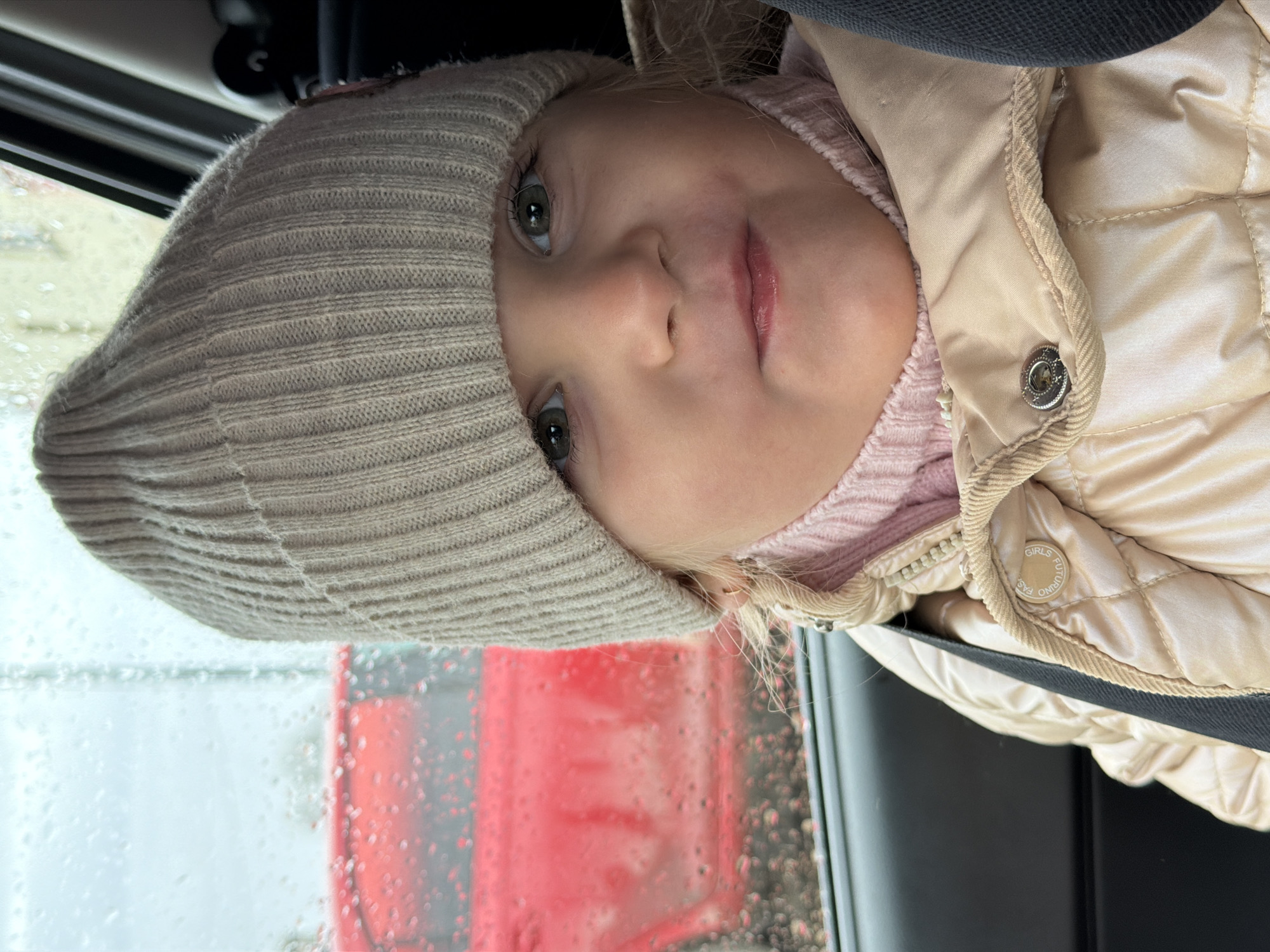 A young child wearing a beige knit hat and a tan puffer jacket is looking at the camera. The child is in what appears to be a vehicle, with a rain-streaked window behind them. The reflection of a red vehicle is visible in the window. Without additional information, it's impossible to confirm if the location is in the city of Chaikovsky, Russia.