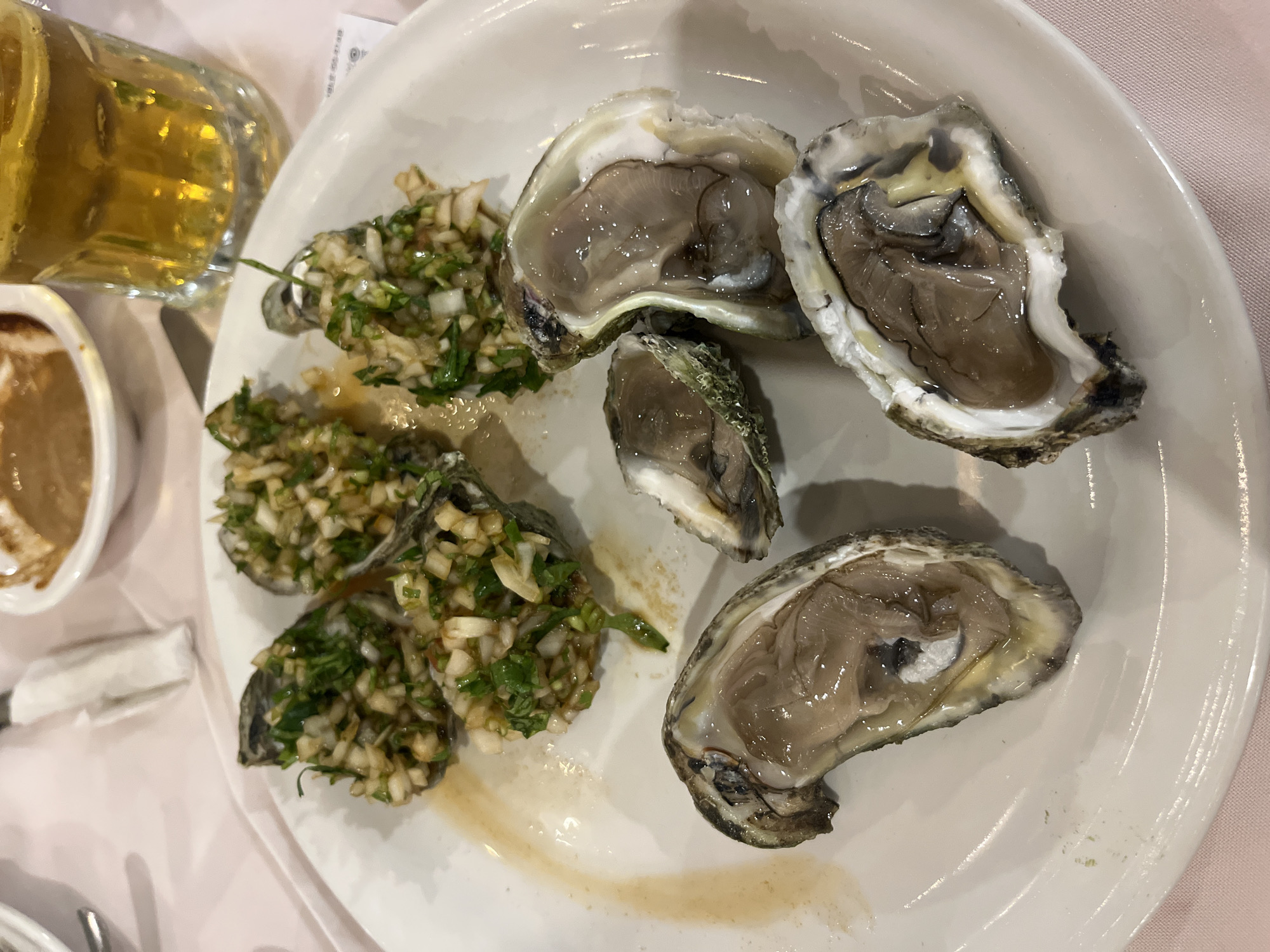 A white plate holds four open oysters on the half shell, topped with a chopped mixture of white and green ingredients, positioned on the left side of the plate. To the right of these are three more open oysters on the half shell, without any topping. The plate is placed on a pale pink tablecloth. In the upper left corner is a glass filled with a yellow liquid, and partially visible is a white container with a brown sauce. The setting appears to be indoors, likely a restaurant, with the location inferred to be San Bernardino Tlaxcalancingo, Mexico. No people are visible in the image.