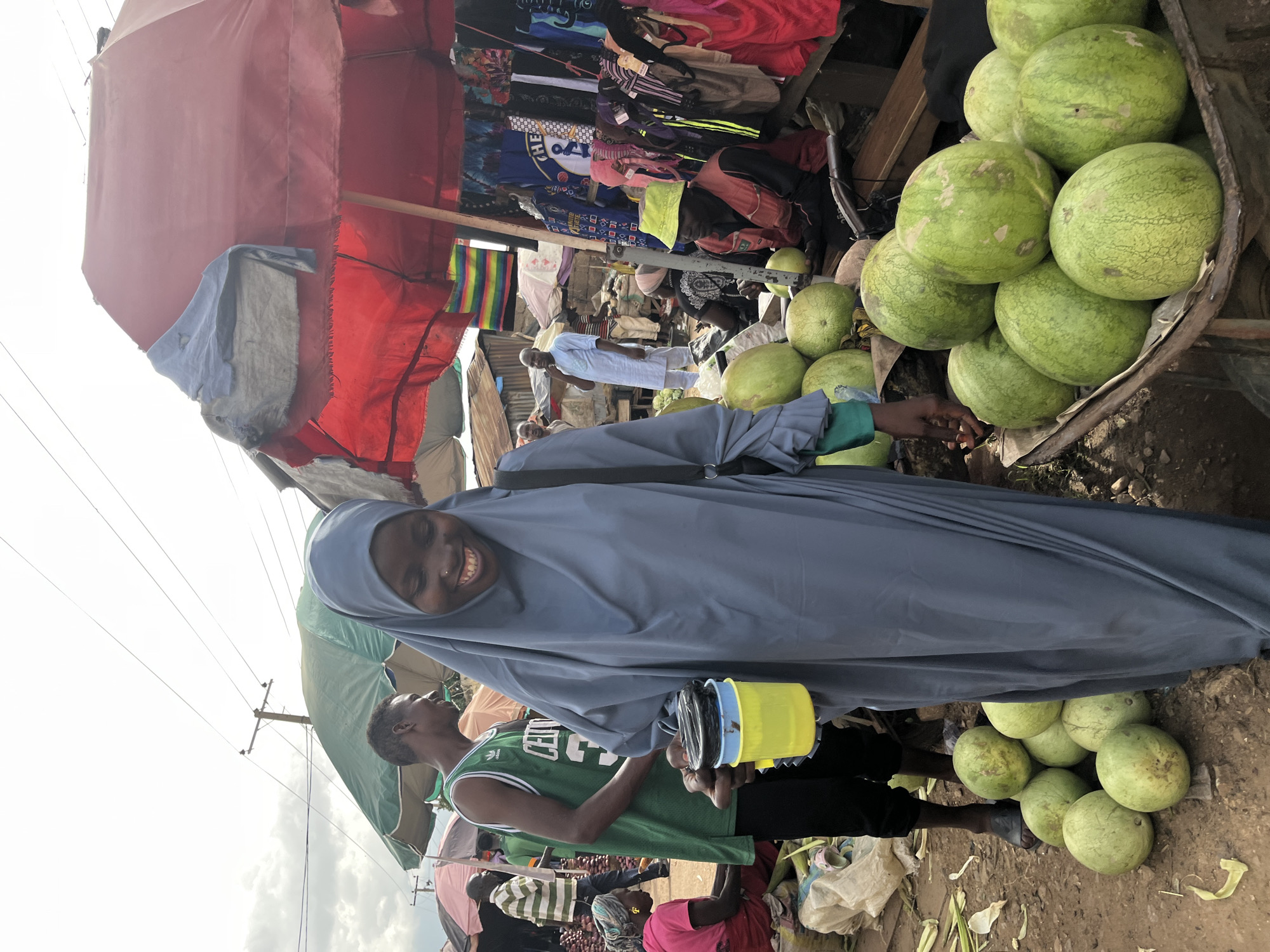 The image depicts an outdoor market scene in Rafin Par, Nigeria. In the foreground, a woman wearing a grey hijab and a blue abaya is smiling and holding a yellow container with black items inside. To her left, a man wearing a green basketball jersey stands beside her, also looking towards the right. Behind them, the market bustles with activity. Various stalls are visible, covered by colorful umbrellas and awnings. One stall displays clothing and textiles, including items with a rainbow pattern and shirts with logos. Another stall features a pile of watermelons in a cart, and several more watermelons are scattered on the ground. Other individuals are seen in the background, including a person in a light blue traditional outfit and another person in a green and white striped shirt. The ground is dirt and scattered with debris. The overall atmosphere suggests a vibrant and busy marketplace.