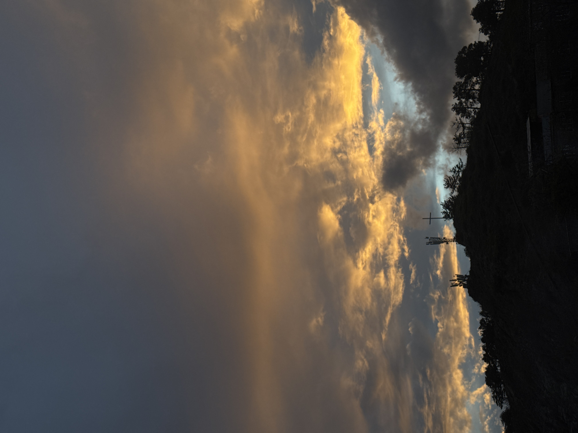 The image, oriented horizontally, depicts a dramatic sky at either sunrise or sunset, with a silhouetted landscape on the right side. The sky on the left and central parts of the frame is filled with dynamic cloud formations, predominantly illuminated by a warm, golden light. These golden clouds contrast sharply with darker, ominous clouds and patches of deep blue-grey sky visible through openings. Some areas show faint light rays breaking through the cloud cover.
On the right, a dark silhouette of a hill or mountain range dominates, extending from the bottom to the top of the frame. The ridgeline is adorned with the dark forms of numerous trees, likely conifers. Several structures are discernible within this silhouette: a prominent cross-like mast or tower (possibly a communications tower or electrical pylon) stands on the lower-middle part of the ridgeline. Further up the slope, indistinct shapes suggest additional buildings or infrastructure, including a rectangular structure with a lighter-colored section, possibly a roof or side.
The warm, golden hues on the clouds against the dark landscape strongly indicate the transitional lighting of either dusk or dawn. The overall scene conveys a sense of natural grandeur and tranquility. There are no people visible, and no discernible text is present in the image. The setting appears to be an elevated, possibly mountainous or hilly, rural environment.