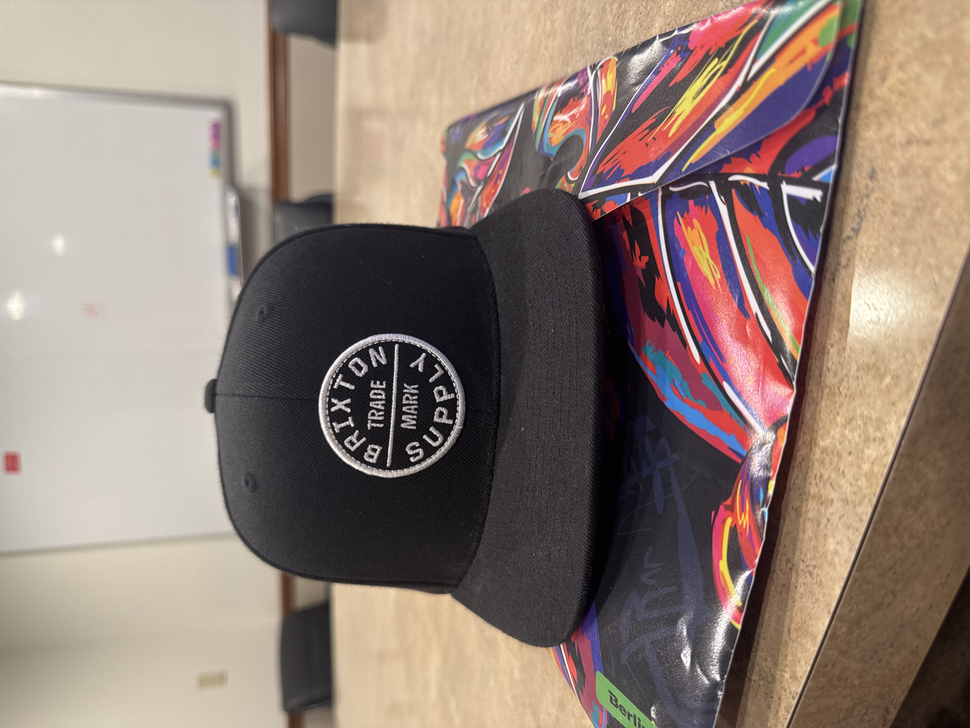 A close-up, rotated shot shows a black snapback hat with a circular logo on the front. The logo is white and embroidered with the words "BRIXTON TRADE MARK SUPPLY" around the circumference, and a "B" in the center. The hat is placed on top of a colorful, abstract printed surface. The surface appears to be paper or cardstock with a dynamic design of broad brushstrokes in red, yellow, blue, and orange against a black background. This printed material is partially folded. In the background, a whiteboard is visible, with some faint markers or stains on it. A wooden table surface fills the lower part of the frame and extends into the background. The overall setting appears to be an indoor space, possibly an office or a room with a whiteboard. There is no indication of the time of day or weather. The composition is dominated by the hat and the vibrant printed surface.