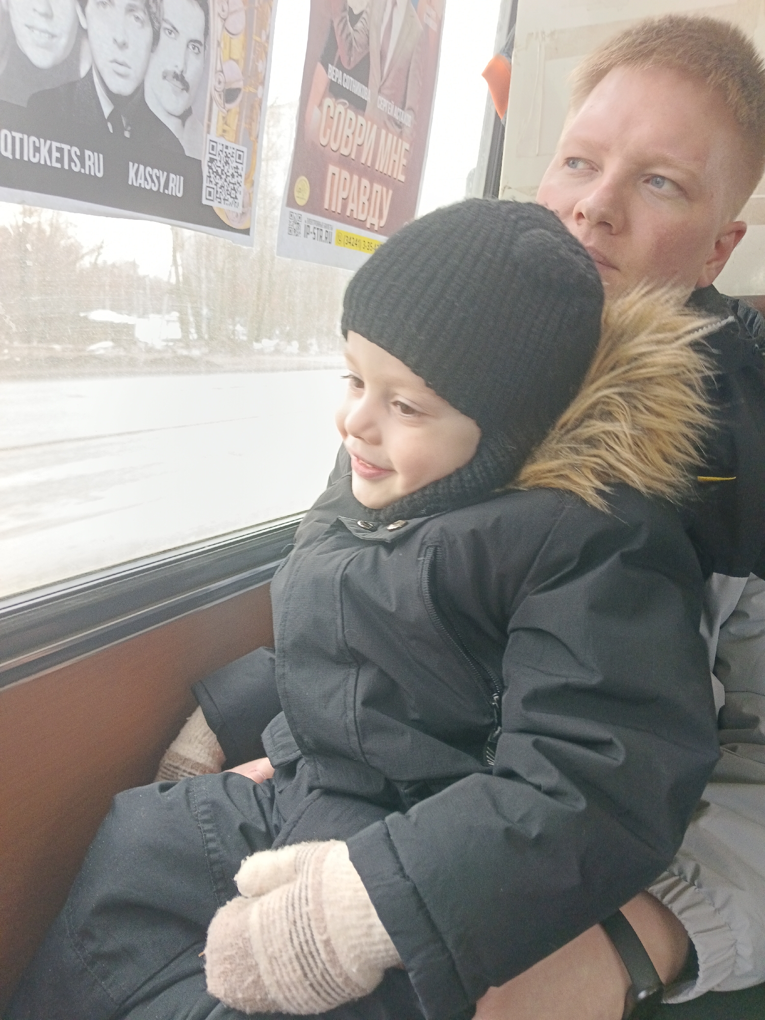 An adult male holds a young child on his lap inside what appears to be a public transport vehicle, both looking out a window at a winter landscape. The male, with short blonde hair and blue eyes, is partially visible on the right, wearing a dark jacket. The child, situated on the left, wears a black knit hat, a black winter coat with a brown fur-lined hood, and striped beige mittens, smiling faintly while gazing out the window.
Above the window, two event posters are affixed. The left poster features four male portraits, including one resembling Freddie Mercury, with visible text "QTICKETS.RU" and "KASSY.RU," alongside a QR code. The right, partially obscured poster advertises a performance titled "СОВРИ МНЕ ПРАВДУ" (Lie to Me the Truth), featuring "ВЕРА СОТНИКОВА" (Vera Sotnikova) and "СЕРГЕЙ АСТАХОВ" (Sergey Astakhov). This poster also displays "IP-STR.RU" and a local phone number with the prefix (34241), which corresponds to the city of Chaikovsky, Perm Krai, Russia.
Outside the window, a blurry, muted winter scene shows snow-covered ground and bare trees under an overcast sky. The interior includes a dark brown window sill or frame.