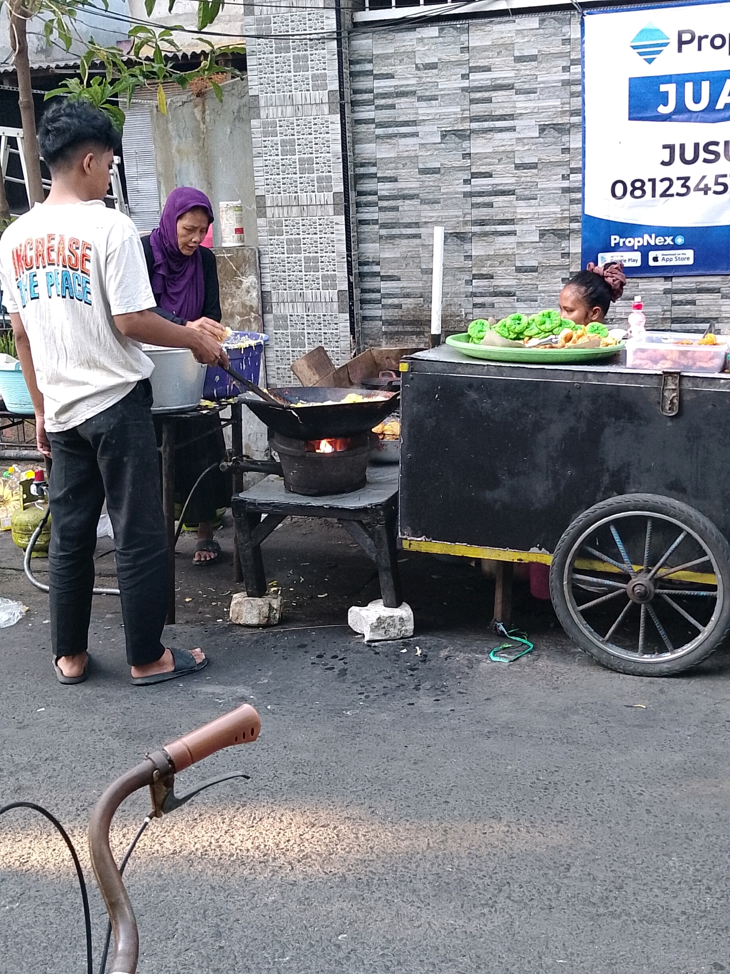 An outdoor scene in Surabaya, Indonesia, depicts a street food vendor and two customers. On the left, a young male wearing a white t-shirt with "INCREASE THE PEACE" printed on it and black pants stands facing a food cart. He appears to be interacting with a woman in a purple hijab and black clothing, who is also positioned near the food cart. The woman is looking down, possibly preparing food.
The food cart is black with yellow trim and a large silver wheel on the right side. On the cart, a green tray holds several portions of food, some wrapped in green paper. Next to it is a clear plastic container with more food items. A woman's head with her hair tied up is visible behind the food items, suggesting she is the vendor.
In the foreground, a portion of a bicycle's handlebars is visible. The background shows a building with a grey, patterned tiled facade. A propane tank is positioned to the left of the scene, and a ladder is leaning against the building. The ground is paved with asphalt. The lighting suggests it is daytime, possibly late afternoon, with shadows cast on the ground.