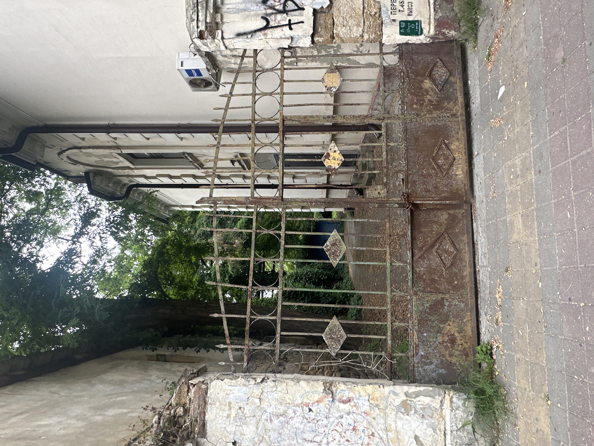 The image shows a rusty, ornate metal gate in front of a building in Stavropol, Russia. The gate is closed and appears to be old, with peeling paint and visible rust. Behind the gate, a pathway leads into a green, overgrown area with trees. To the right of the gate is a section of the building with an air conditioning unit mounted on the wall, and graffiti on the stone pillar. To the left of the gate is a crumbling, textured wall. The ground in front of the gate is paved with interlocking blocks. There are no people visible in the image.