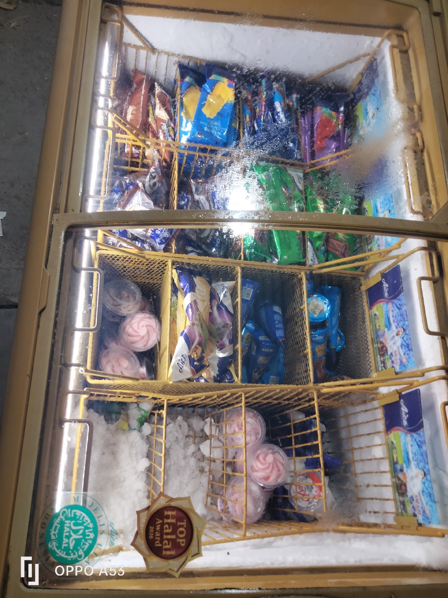 This is a top-down view of a commercial chest freezer, likely located in Karawang, Indonesia. The freezer is filled with various frozen treats, predominantly ice cream bars and small ice cream cups. The interior is organized with yellow wire shelves, creating compartments for different products. Some compartments contain individually wrapped ice cream bars in colorful packaging, such as blue, yellow, and green. Other compartments hold small, clear plastic cups with pink-swirled ice cream.
The freezer door is transparent, revealing the contents. Condensation and frost are visible on the glass, indicating the cold temperature. In the lower left corner of the image, there are two certification stickers: one round green sticker with Arabic calligraphy and the word "HALAL" in English, and a hexagonal brown and gold sticker that reads "TOP Halal Award 2026". The bottom left of the image also shows the text "OPPO A53", which is likely a watermark from the device used to take the photo. The overall environment appears to be a commercial establishment selling frozen goods.