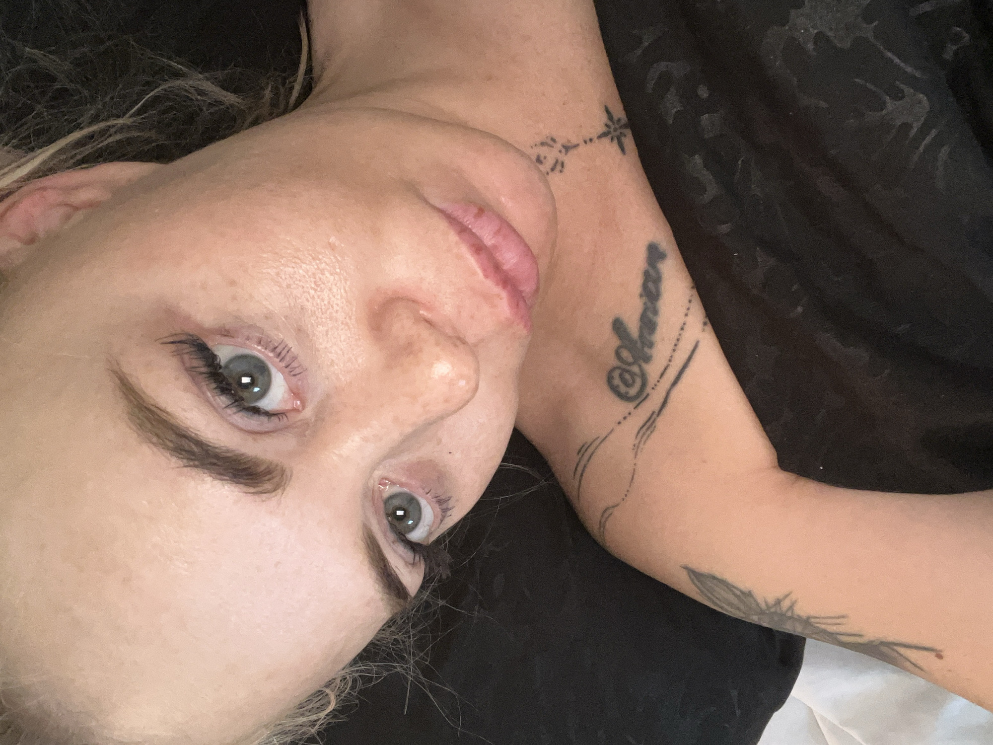 A close-up, high-angle, indoor selfie shows a fair-skinned person with blonde hair and light blue eyes. They are wearing black fabric, possibly a shirt or blanket, which covers their chest and shoulder. Tattoos are visible on the person's shoulder and neck. The person's face is partially obscured by the framing of the selfie. The background is dark and out of focus.