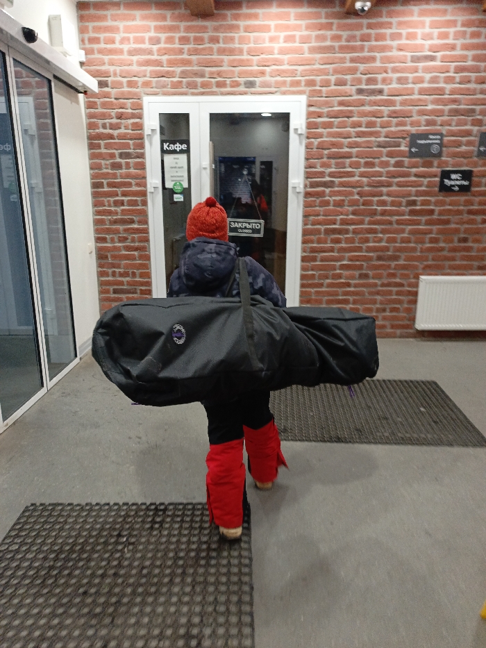 The image captures a person, likely a child, seen from behind, walking through what appears to be an indoor or enclosed entryway of a building. The person is dressed in winter attire, consisting of a red knitted hat with a pom-pom, a dark winter jacket (possibly with a camouflage pattern), black pants, and bright red leg warmers or gaiters worn over their shoes. They are carrying a large, black, horizontally oriented duffel-style bag over their back, supported by a shoulder strap. The bag has a round, possibly purple and white, logo on its side.
The setting features a red brick wall on the right side and in the background, contrasting with white-framed glass doors. To the left, there is another set of glass doors with white frames, reflecting indoor light and some indistinct shapes. In the center of the background, a set of double white-framed glass doors leads into what is identified as a "Кафе" (Cafe) by a sign on the left door. Another sign on the right door of this set clearly indicates "ЗАКРЫТО" (Closed) in red letters on a white background.
Above the central doors, a security camera or sensor is mounted on the ceiling. On the brick wall to the right of the central doors, there are two small black directional signs. The upper sign has an arrow pointing left with some unreadable text. The lower sign clearly indicates "WC Туалеты" (WC Toilets) with an arrow pointing right. Further to the right on the brick wall, a white radiator is visible.
The floor is a light grey, possibly concrete or tiled surface, with two rectangular dark, textured floor mats placed strategically. The person is walking from the foreground towards the central, closed cafe doors, creating a sense of movement or transition. The lighting suggests an indoor artificial light source. The overall impression is that of someone at a recreational facility, possibly a ski resort or a winter sports complex, given the attire and the size/type of the bag, which could hold skis or a snowboard.