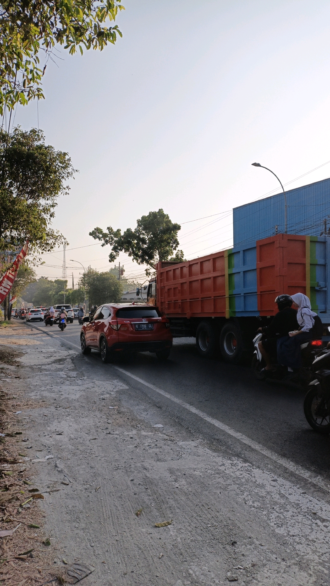 The image depicts a traffic scene on a moderately busy road, likely in an urban or semi-urban area of Sumbring, Indonesia. The time of day appears to be late afternoon or early morning, indicated by the warm, low-angled sunlight casting long shadows and illuminating the sky with a hazy, light blue hue.
Dominating the right side of the frame is a large, multi-colored truck. The truck's bed is bright orange, and its cab and trailer section are segmented with bands of blue and yellow. It appears to be stationary or moving very slowly, as is the traffic around it.
In front of the truck, and slightly to the left, is a red SUV, also in traffic. The SUV has a license plate that reads "K 1709 GL". Further to the left and in the background, several motorcycles and other vehicles are visible, indicating moderate traffic congestion. One motorcycle in the foreground, on the right side of the image, carries two passengers, a driver and a passenger wearing a white hijab and dark clothing.
The road surface is asphalt, with a white line marking a lane divider visible. The left side of the road features a dusty, uneven shoulder with scattered debris, fallen leaves, and some vegetation. Trees with green foliage are present on both sides of the road, with some taller trees and a distant tower visible in the background. A red and white flag or banner is attached to a pole on the far left side of the image. The overall impression is a typical, everyday traffic situation in a developing Indonesian city.