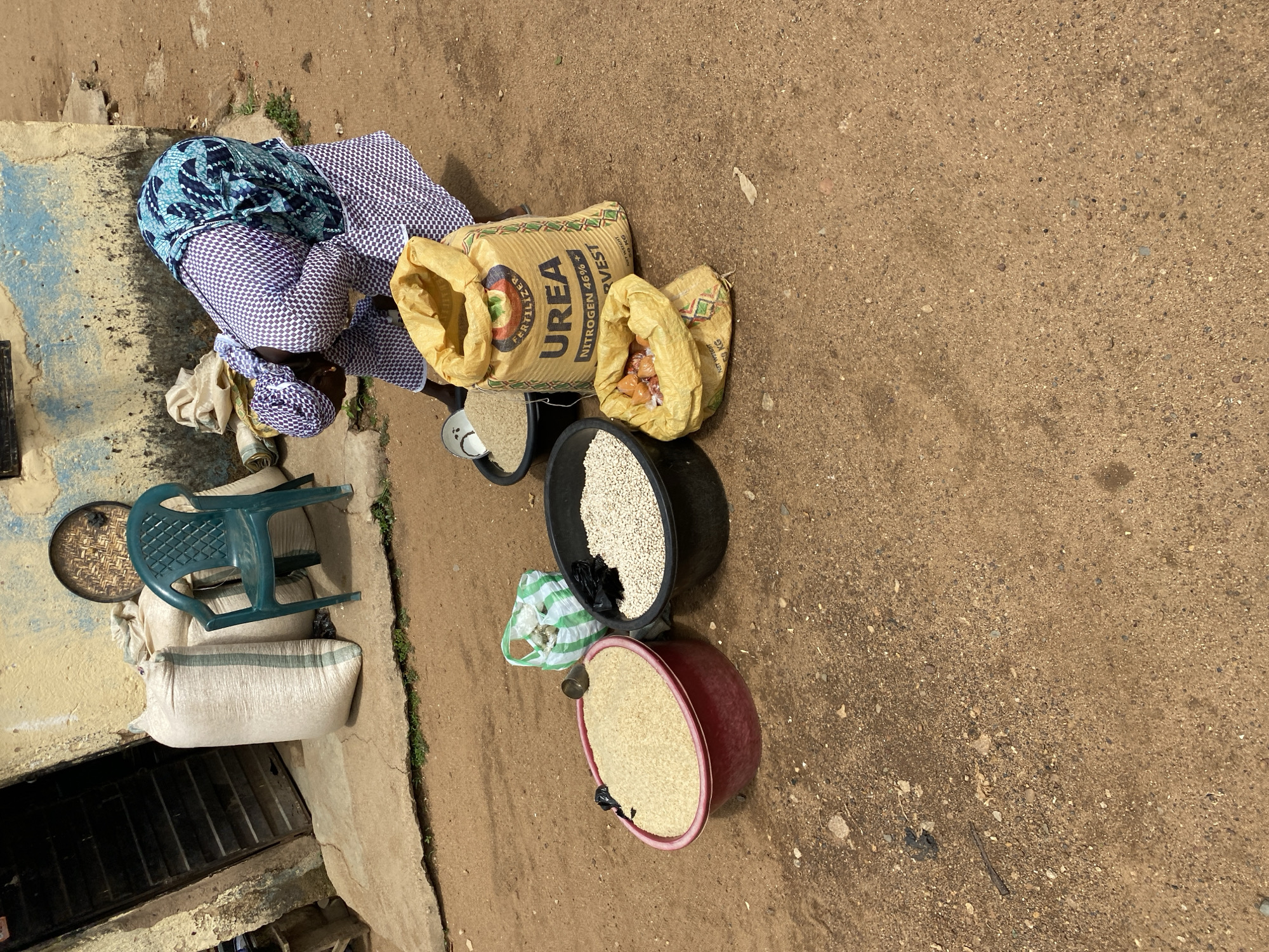 A woman dressed in a blue and white patterned outfit is bent over near several bags and containers filled with goods in Bukuru, Nigeria. A large bag labeled "UREA" is visible. There are also bags containing what appear to be small packaged items. Baskets filled with grains or beans are near the urea. A green plastic chair is stacked on top of a large white bag near a building with a partially visible doorway. The ground is a sandy, dirt surface.