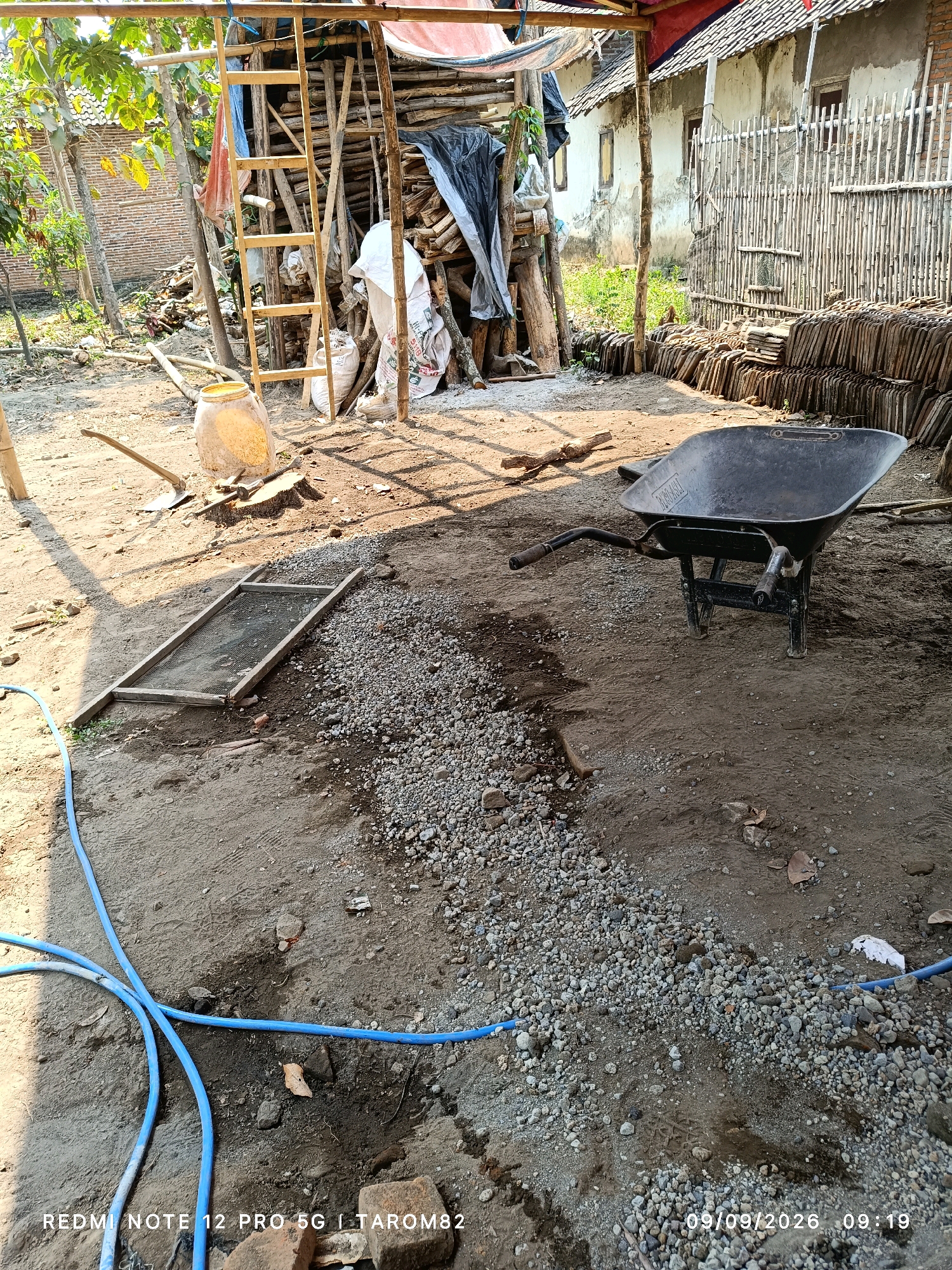 The image depicts a construction site in Rejoagung, Indonesia, characterized by an outdoor setting under a makeshift shelter. In the foreground, a black wheelbarrow rests on the dusty ground, which is partially covered with small stones and gravel. A blue hose snakes across the scene, connected to a water source. To the left, a wooden sieve or screen is laid flat on the ground. Further back, a tall wooden ladder leans against a structure made of bamboo and wood, which appears to be supporting roofing materials. Piles of lumber, some covered with tarpaulin, are visible. A large yellow plastic drum sits near the ladder. In the background, a rustic house with a tiled roof stands adjacent to a bamboo fence. Stacks of roof tiles are neatly arranged to the right of the house. The environment suggests ongoing construction or renovation work. The overall scene is one of a practical, outdoor workspace with construction materials and tools.