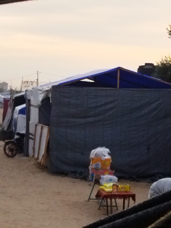 The image displays an outdoor scene in what appears to be a temporary settlement or camp within the city of Khan Yunis, Palestinian Territory. The ground is composed of light brown sand or dirt. The sky is a pale, hazy yellow-white.
A series of makeshift structures, predominantly tents or shelters constructed from tarpaulins and other materials, line the midground. The structure on the immediate right features dark gray tarpaulin walls and a blue tarpaulin roof, supported by visible wooden beams, forming a peaked shape with an open section near its apex. To its left, additional shelters are visible, constructed from white and blue tarps, with some white panels leaning against one. A bicycle with a cart or utility attachment is parked near the leftmost visible shelter.
In front of the dark gray structure, two small, makeshift display tables are present. The taller table on the left consists of a green metal frame holding stacked boxes, topped with items contained in a white plastic bag. To its right, a shorter table draped with a reddish cloth displays various packaged goods.
In the background, indistinct outlines of distant structures, utility poles, and wires are visible. A dark water tank is discernible on the roofline of a far-off building. No individuals are visible in the frame.