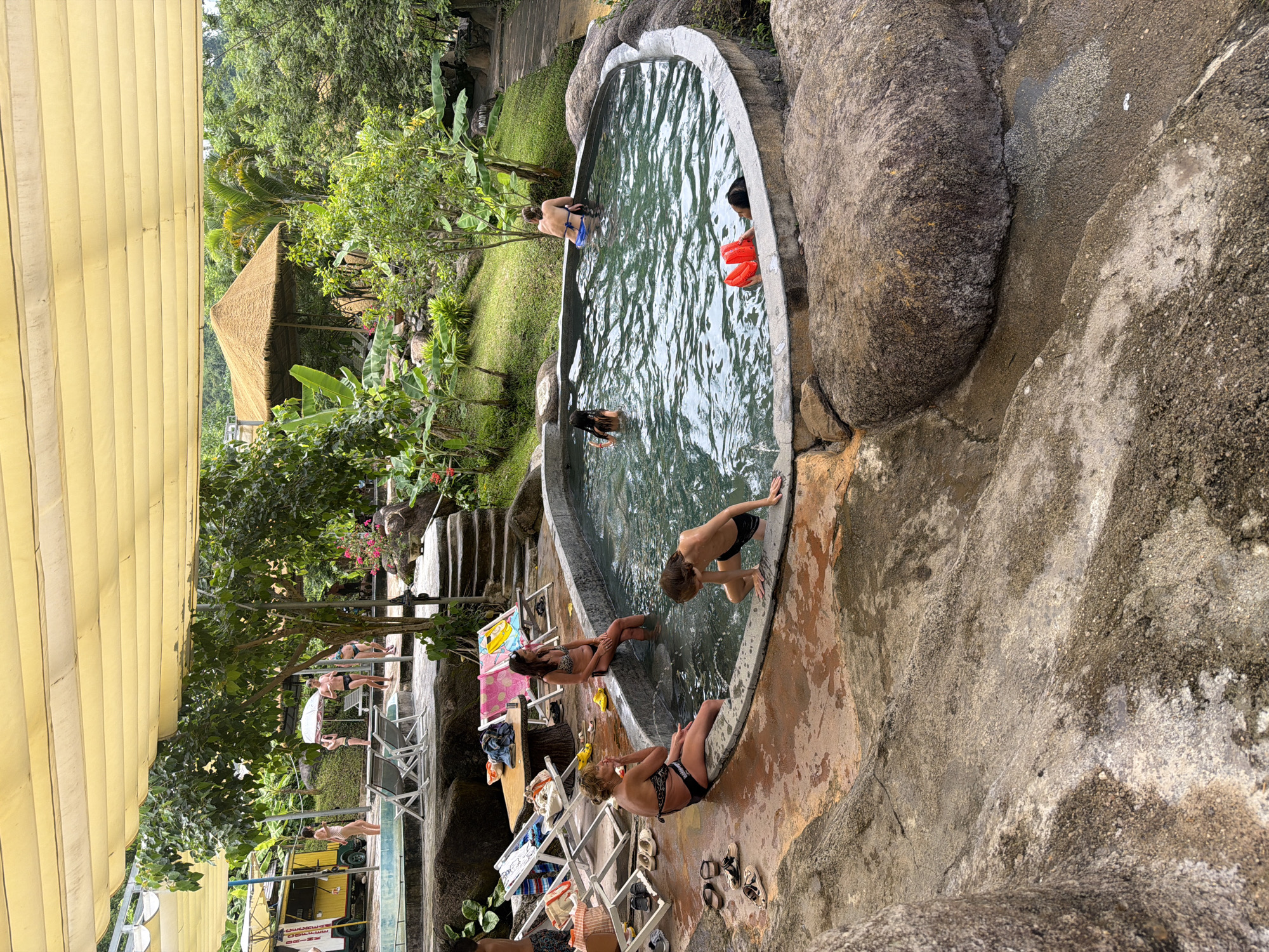 The image depicts an outdoor hot spring resort nestled within a lush, tropical environment. In the foreground, a circular, stone-lined hot spring pool is filled with rippling, clear water. Several individuals are visible in and around the pool. One woman, wearing a black and white bikini, sits on the edge of the pool, facing left, with her hand on her chin. A young boy in black swim trunks kneels at the edge of the pool, reaching into the water with his left hand. Further into the pool, a person with dark hair, wearing a dark bikini, is partially submerged. On the opposite side of the pool, a person wearing a blue bikini bottom and a dark top is leaning against the edge. Two red inflatable armbands are floating near the surface.
Surrounding the pool is a natural, rocky terrain with patches of wet, reddish-brown concrete. Several lounge chairs are set up on this area, some with folded towels and bags. To the left of the pool, a yellow food cart with an awning is visible, and two women in swimwear stand near it.
The background features a dense green landscape with mature trees, including banana plants, and a thatched-roof structure. A paved pathway can be seen winding up a grassy slope in the distance. The entire scene is covered by a large, pale yellow awning supported by poles. The resort appears to be located in the city of Khanh Vinh, Vietnam, given the tropical vegetation and architectural style.
