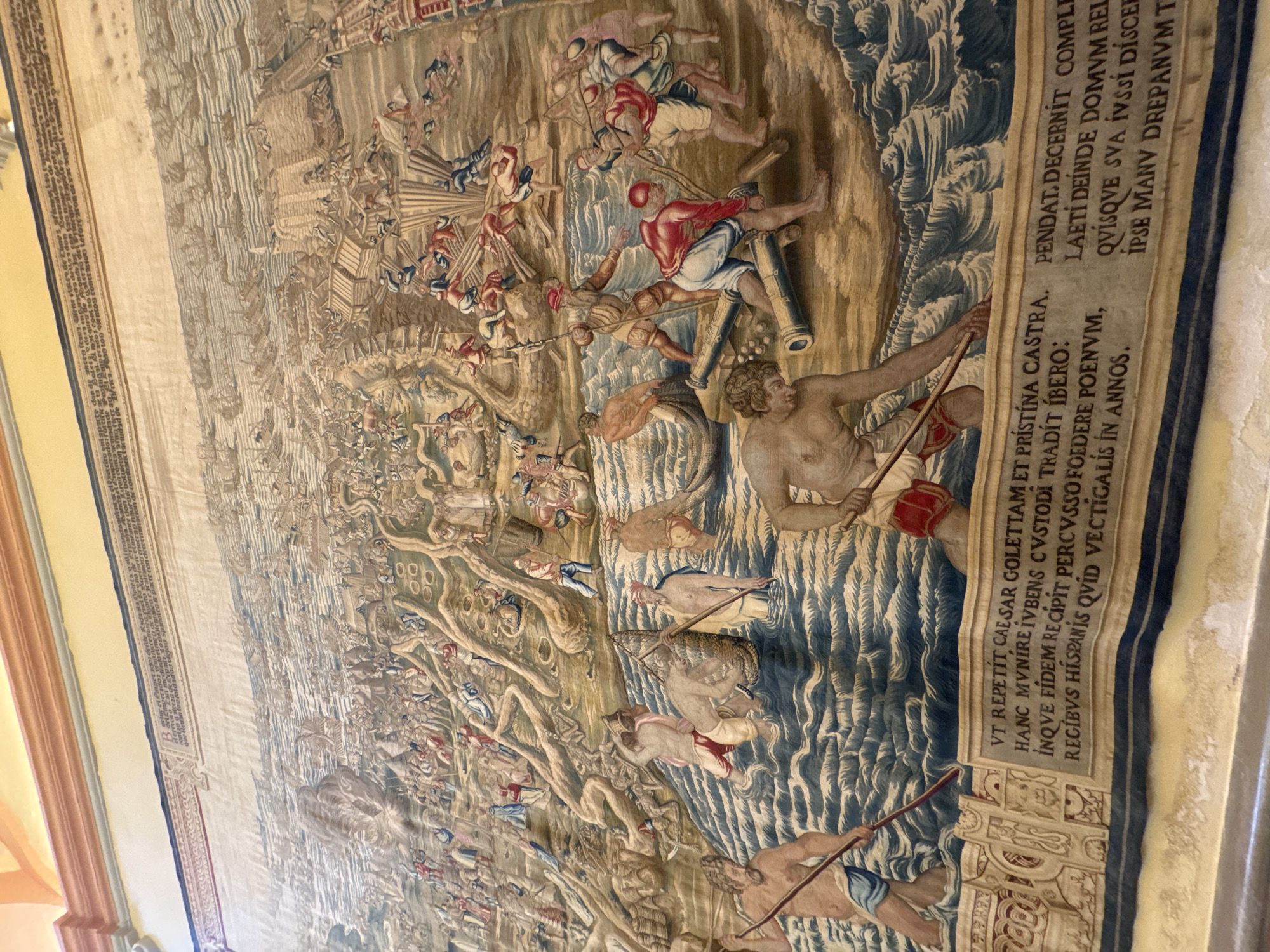 The image depicts a large, detailed tapestry hanging on a wall, likely in a historic building in Seville, Spain. The tapestry illustrates a complex historical or mythological scene involving numerous figures engaged in battle and maritime activities.
In the foreground, several male figures are depicted in the water. One man, with a beard, stands with a spear. Another, muscular figure is shown working in the water, and further right, a group of men are handling cannons and spears, some wearing red garments. In the background, a large fleet of ships is visible on the sea, with many smaller figures engaged in various actions, including construction of wooden structures, combat, and movement across land and water. The scene appears chaotic, suggesting a significant event like a battle or naval engagement.
Below the main scene, a decorative border runs across the bottom of the tapestry, featuring intricate carvings. A prominent band of text is present within this border, containing Latin inscriptions. These inscriptions are likely to provide context or a narrative for the depicted scene. The tapestry's colors are muted and aged, typical of historical textiles, with dominant tones of blue for the water and earthy browns and beige for the land and figures.
The ceiling visible at the top of the image appears to be ornate, suggesting the tapestry is displayed in a significant and well-decorated setting, consistent with a historical site in Seville such as the Alcázar or a cathedral.