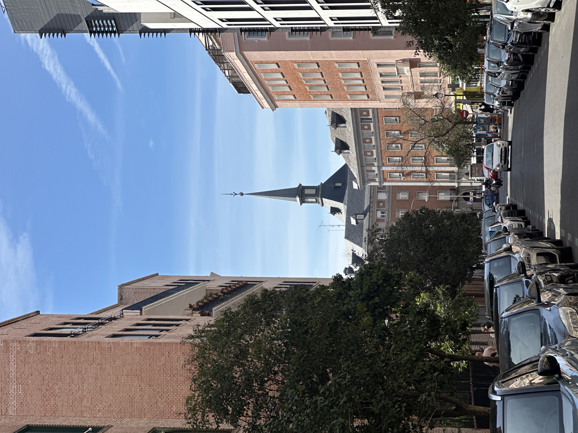 The image captures an urban street scene in Madrid, Spain, looking up between buildings towards a clear blue sky. In the center, a tall, narrow church spire rises above the rooftops. On either side, buildings of varying architectural styles line the street, with facades of brick and stucco, and visible windows and balconies. The street is flanked by rows of parked cars on both sides, and green trees provide a natural element to the urban landscape. The scene suggests a sunny, daytime environment with a mix of residential and potentially commercial structures. A few people are visible in the distance, walking on the sidewalk.