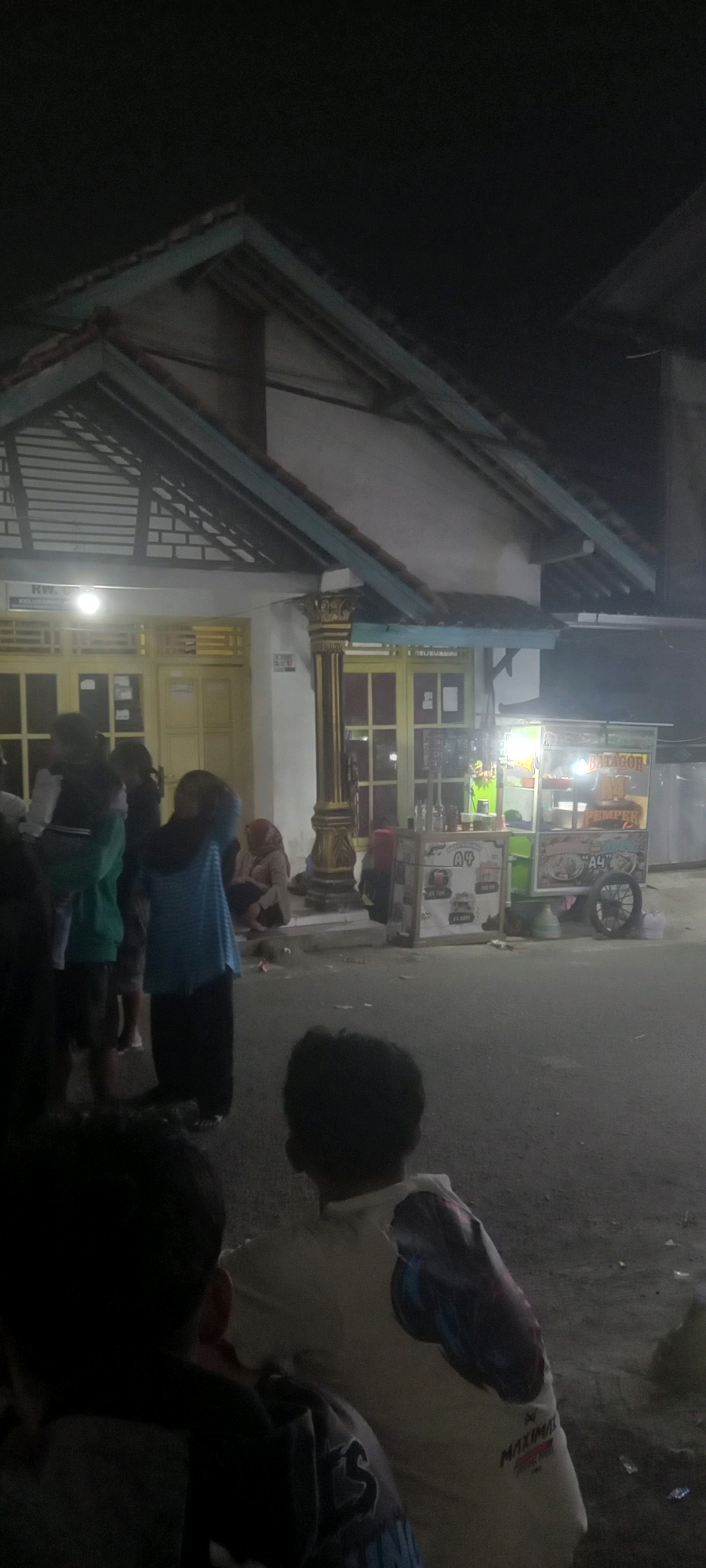 The image shows a street scene at night in Argasoka, Indonesia, with a food cart prominently displayed. Several people are visible, some standing and others sitting. A group of individuals are gathered near the food cart, which is illuminated by bright lights, suggesting it is operational. The cart appears to be selling food items, with visible signage. In the foreground, two children are seen from behind, facing away from the camera. The background shows a building with yellow-tinted windows and decorative pillars. The overall atmosphere is that of a lively evening street environment.
