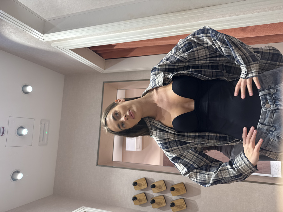 A female subject is centrally positioned within what appears to be a retail store fitting room, located in Verona, Italy. She has long, dark brown hair and fair skin, with visible facial makeup. The subject is wearing a black, deep V-neck strapless bodysuit layered under an unbuttoned, long-sleeved plaid shirt with blue, white, and beige patterns. Light blue denim jeans are visible below her waist. Her hands are placed on her hips, thumbs forward. Her gaze is directed upwards and slightly to the viewer's left, with a neutral expression.
The room features muted pink-beige textured walls. The white ceiling contains three recessed circular and square downlights, along with a square-shaped fixture displaying an illuminated green light. To the left, a light-framed doorway opens into an adjacent room with matching walls and a white wall-mounted air conditioning unit. Below this doorway, six rectangular wooden plaques, each fitted with a black hook, are affixed to the wall in two vertical rows of three. To the right, another light-framed doorway reveals a reddish-brown curtain within, indicating a separate changing area. The floor is not visible.