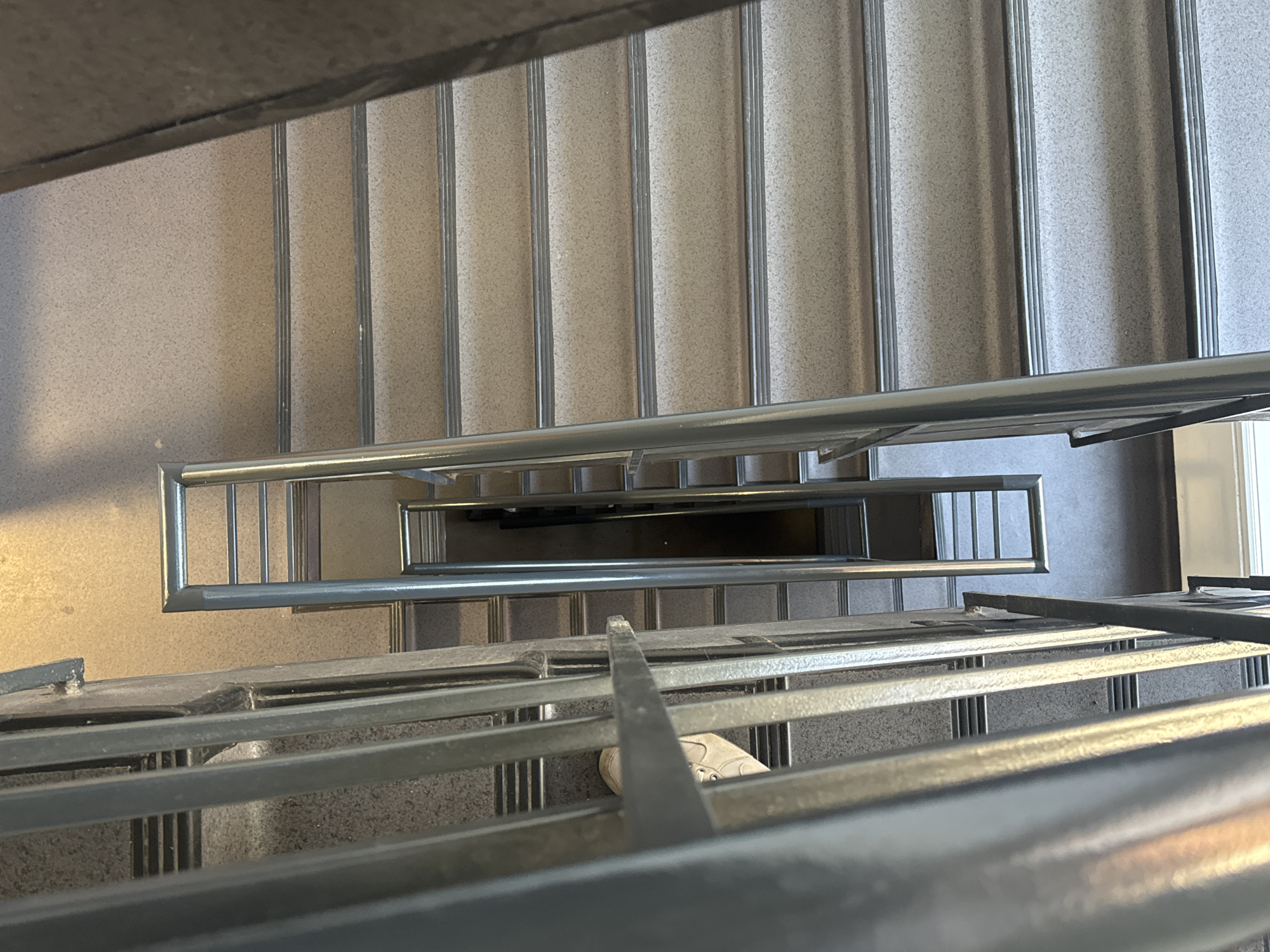 The image shows a downward view of a staircase with metal railings. The stairs are covered with a light-colored, speckled material. A single foot wearing a white sneaker is visible on one of the steps. The staircase appears to be indoors, likely in a public building. The location is unknown and cannot be determined based on the image.