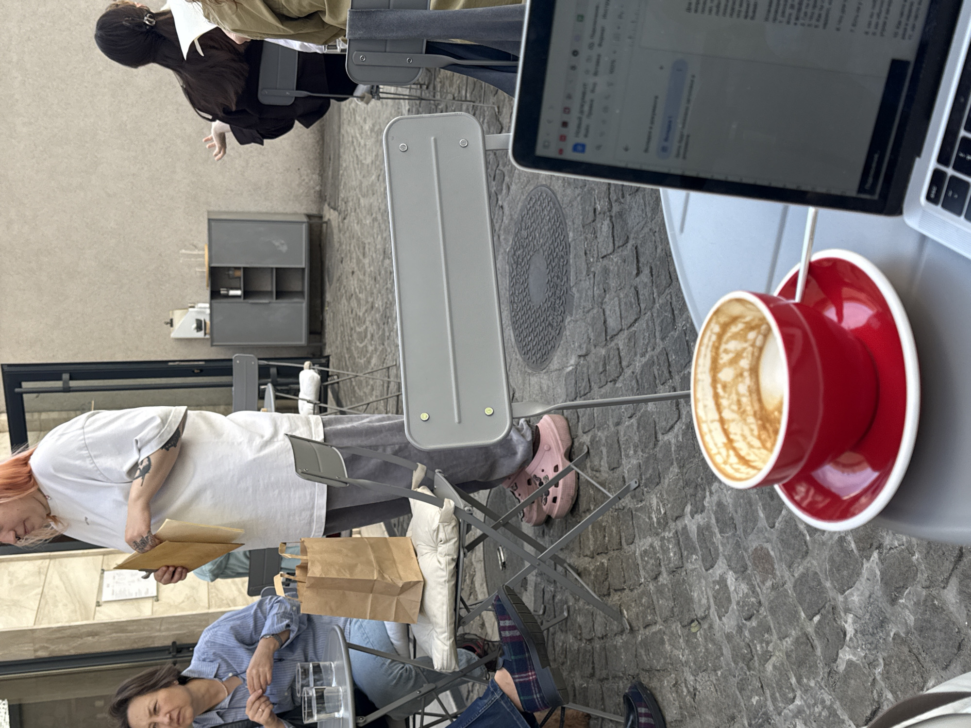 This outdoor scene shows three people seated at tables on a cobblestone street, presumably in Vracar Urban Municipality, Serbia. The focus is on a laptop and a red coffee cup placed on a table. A person with pink footwear sits adjacent to another person in a striped shirt and jeans, and an opened paper bag. Another person is also visible in the background.