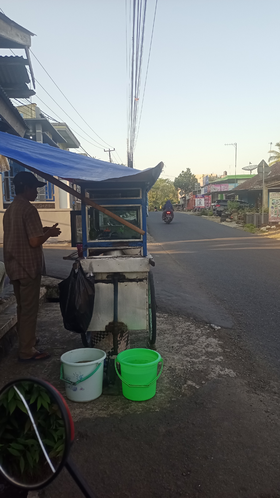 A street vendor stands next to a food cart in Karyamandala, Indonesia. The vendor, wearing a checkered brown shirt and dark pants, is holding a small object in their hands. The food cart, with a blue tarp roof and metal framing, displays food items within a glass enclosure. Two buckets, one white with a floral pattern and the other bright green, are placed on the ground next to the cart. A black plastic bag hangs from the side of the cart. In the background, a motorcycle is seen driving away on a paved road lined with buildings and trees. The sky is clear and pale blue, suggesting early morning or late afternoon light. The image is taken from a low angle, possibly from the perspective of someone on a motorcycle, as evidenced by the reflection in a circular mirror visible in the lower left corner.