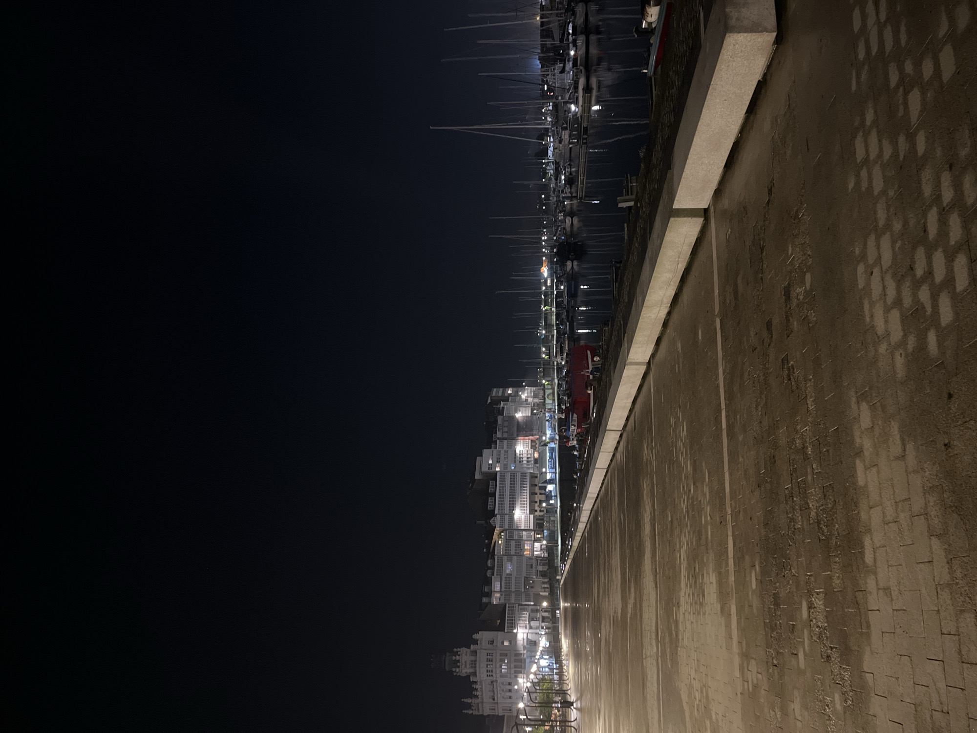 This image depicts a nighttime scene in A Coruña, Spain, taken from the promenade alongside the marina. On the right side of the image is a concrete wall with textured paving, suggesting a walkway. Adjacent to the wall, further into the image, is the marina, filled with numerous sailboats, their masts reaching upwards. Across the marina are buildings illuminated at night, some with white balconies. The left portion of the image is predominantly dark, indicating the nighttime sky. No people are visible in the image.