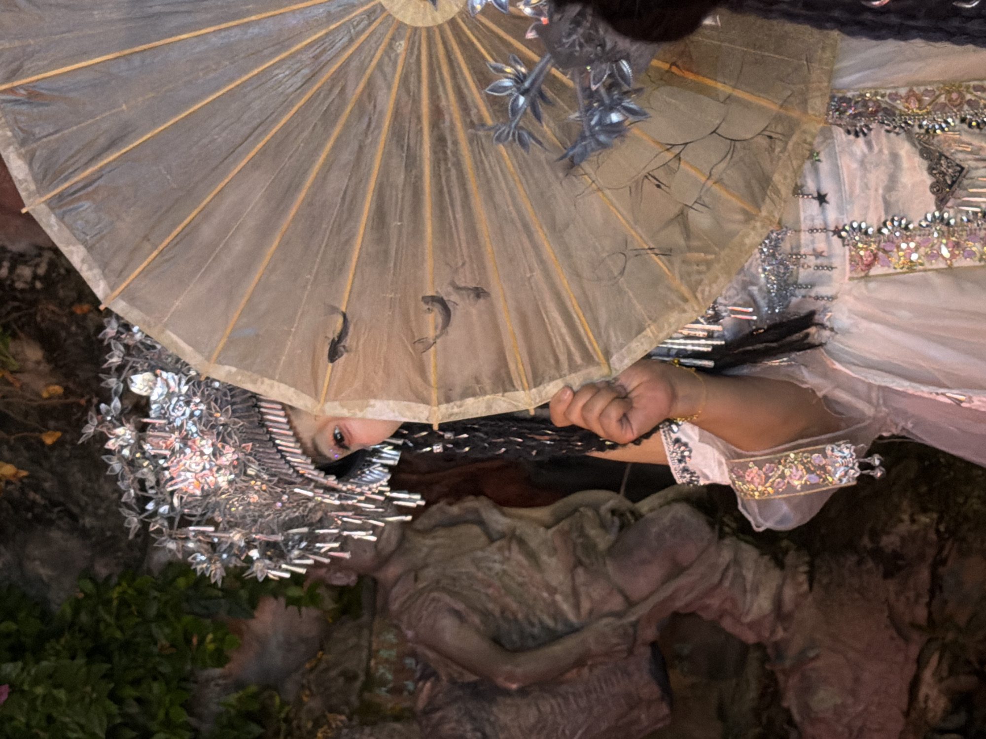 A person in elaborate costume, holding a decorative umbrella, is partially visible in front of a sculpted figure and lush greenery, suggesting an outdoor setting in Yuzhong District, China, possibly a tourist attraction or cultural performance space. The person is wearing a silver headdress with hanging ornaments, intricate beaded jewelry on their wrists and garment, and a white dress adorned with colorful embroidered trim and more hanging embellishments. The umbrella is translucent with wooden ribs and features painted depictions of fish and branches. The sculpted figure, to the left, appears to be a male statue in rough-hewn clothing. The background consists of dark foliage and textured rock formations.