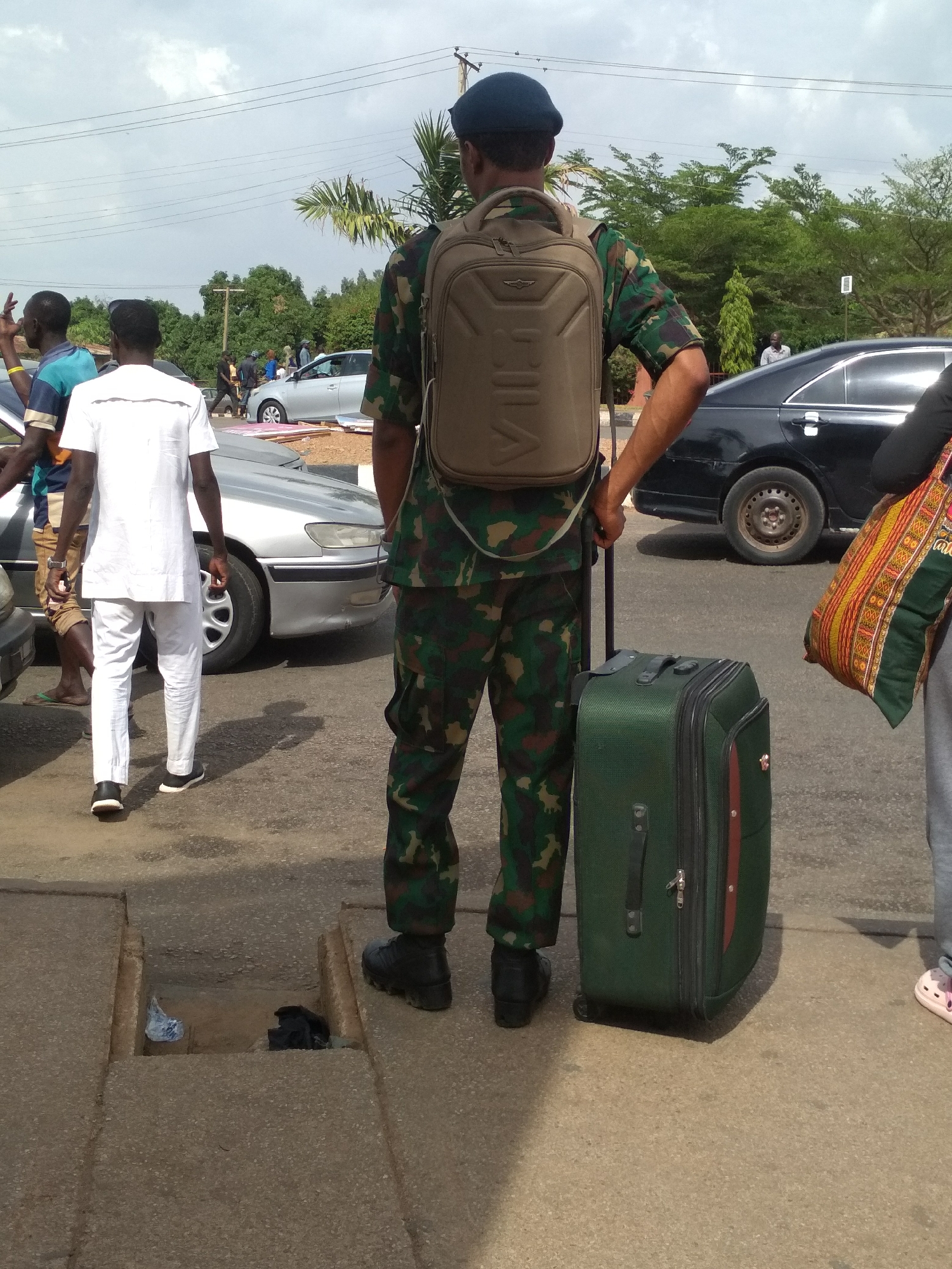The image depicts an outdoor street scene in Barakin Lerawi, Nigeria, on an overcast day.
The primary subject is a man seen from behind, wearing a military-style camouflage uniform, a blue beret, and black boots. He carries a tan backpack with the word "VIVE" faintly visible on its hard shell. He is holding the extended handle of a dark green wheeled suitcase, which has a prominent red stripe on its side.
To his left, another man in a white tunic and trousers walks away from the camera. Further left, a man in a striped blue and brown top is visible, with his arm raised in a gesture. Partial view of a person's legs and a foot in pink slippers is seen on the far right, next to the man in uniform. Several other individuals are visible in the background, some walking, some standing near vehicles.
The scene is set on an asphalt road with a concrete pedestrian area. Various cars, including a silver sedan and a black sedan, are present, suggesting a busy street or a parking area. In the foreground, an open concrete drainage channel containing some litter (a plastic bag and a dark object) is visible on the ground. The background features green trees and palm trees, and overhead power lines traverse the sky.
The activity suggests people are either traveling, waiting for transportation, or moving through a public urban or semi-urban area. The presence of luggage strongly indicates travel. The time of day is daytime, under a cloudy sky.