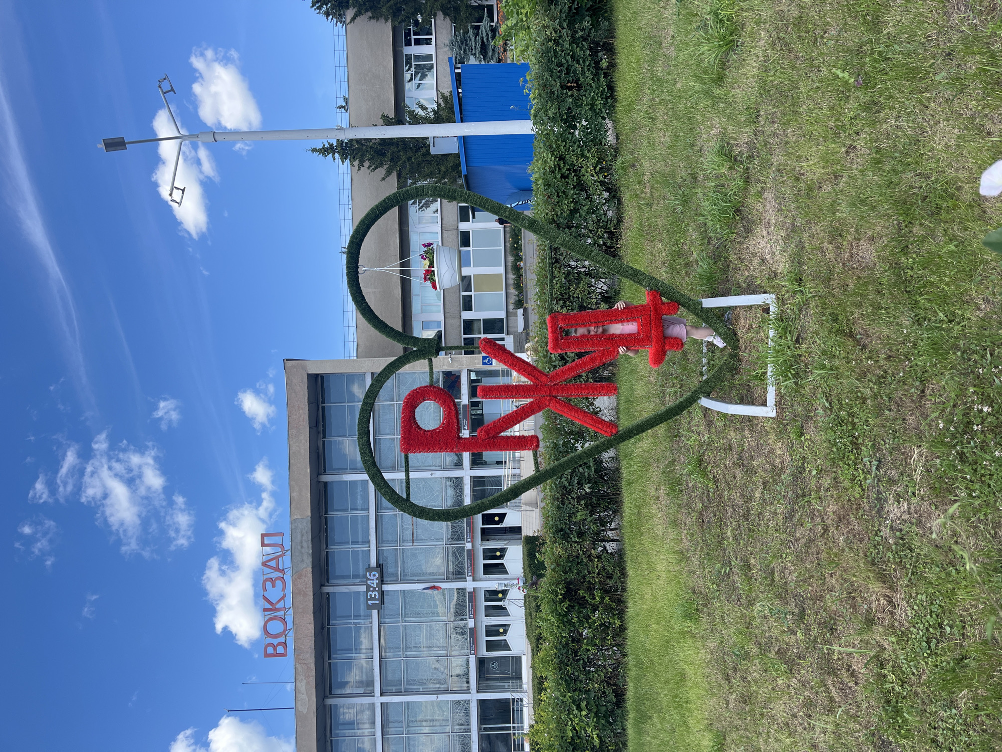 A young child is playing near a large, heart-shaped floral display outside a building in Shadrinsk, Russia. The child is lying on their back, with their head and torso inside the heart and their legs sticking out. They are wearing a pink outfit. The heart-shaped display is made of green foliage and contains large, red, furry letters that spell out "РЖД". The letters are arranged in a way that suggests a decorative element. A white flower pot with pink flowers is hanging from the top of the heart. The building behind the display appears to be a train station, with the word "ВОКЗАЛ" (VOKZAL, meaning train station in Russian) visible in large red letters on its facade. A digital clock on the building shows "13:46". The scene is set on a sunny day with blue skies and scattered white clouds. The ground is covered in green grass, with some areas appearing drier. A street lamp is visible in the upper left corner of the image. The overall atmosphere seems casual and cheerful, with the child enjoying themselves in the decorative outdoor space.