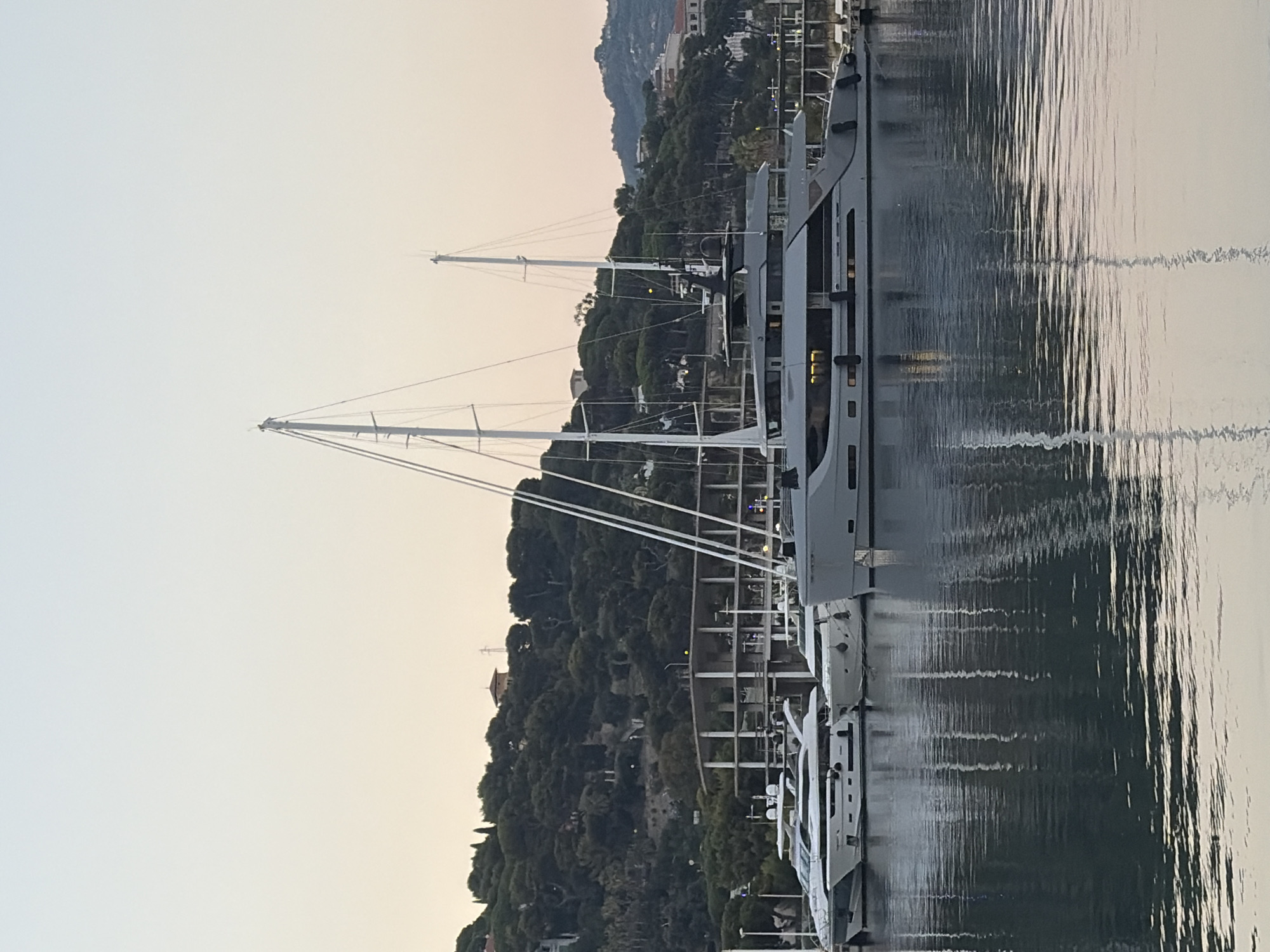 This media file captures a tranquil scene at a marina, likely in Imperia, Italy, as indicated by the location context. The image features several large yachts docked side-by-side. The yachts are modern and sleek, with prominent masts suggesting they are sailing yachts. The scene is set against a backdrop of a lush, green hillside dotted with buildings, hinting at a coastal town. The water in the marina is calm, reflecting the yachts and the sky, creating a slightly distorted, rippling effect.
The time of day appears to be either early morning or late evening, given the soft, diffused light and the muted colors of the sky, which transitions from a pale blue at the top to a warm, peachy hue closer to the horizon. The weather seems to be clear and calm, ideal for a marina setting. There are no people visible in the frame, contributing to the peaceful and serene atmosphere. The focus is on the impressive vessels and the natural beauty of the landscape. The reflections in the water are a notable detail, adding depth and visual interest to the composition. No text is visible in the image.
