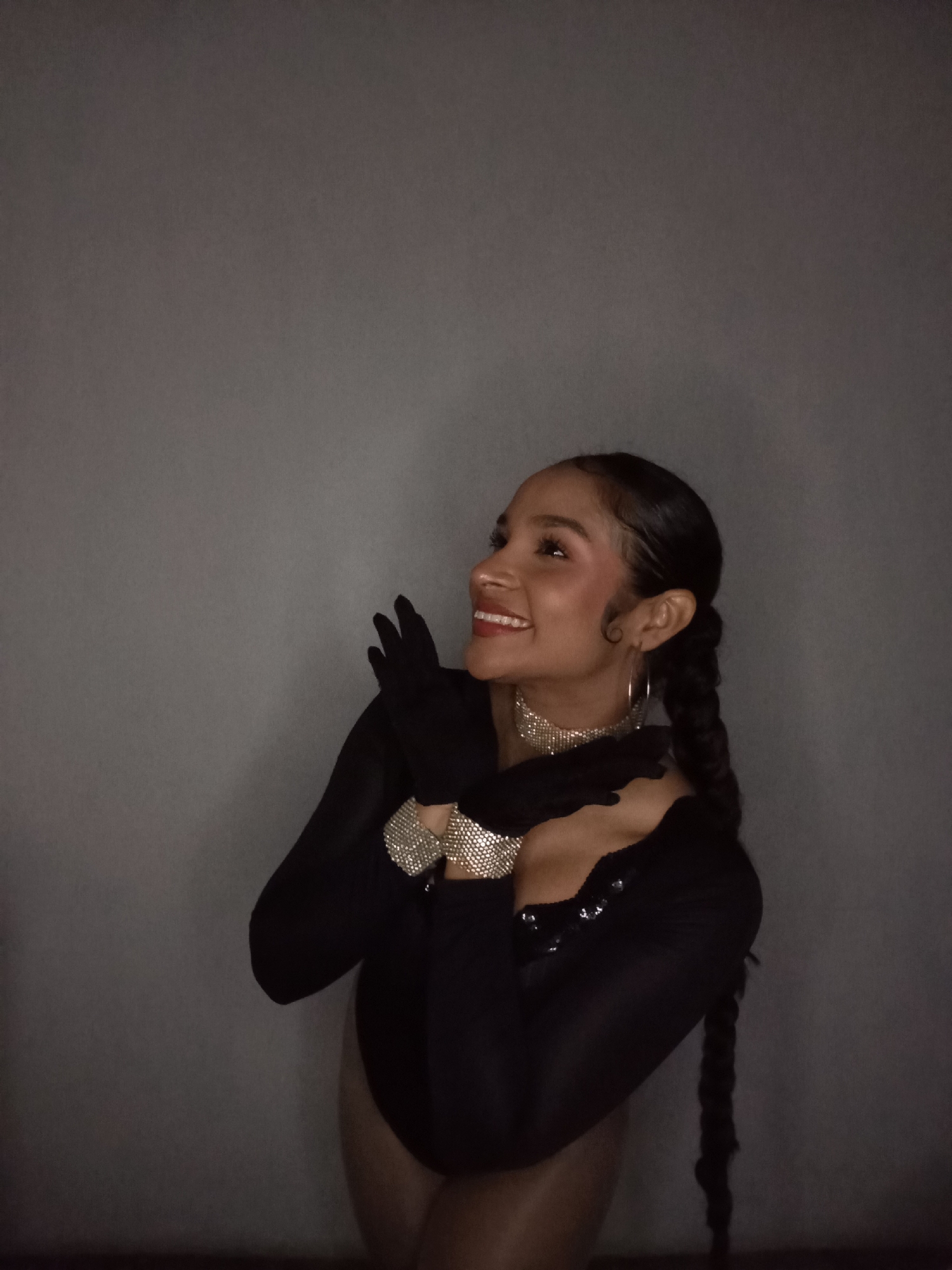 A young woman is captured in a half-body shot, posing against a plain, dark grey wall. She is looking upwards and to her left, displaying a wide, genuine smile that reveals her teeth. Her dark hair is styled into two long, thick braids that rest over her shoulders, with delicate baby hairs framing her face.
She is dressed in a black, long-sleeved bodysuit or leotard, which features subtle sequin embellishments on the chest. Her arms are adorned with long black gloves, and over these, she wears two prominent, sparkling diamond-like cuffs on each wrist. A matching sparkling choker necklace graces her neck, and she wears simple silver hoop earrings. Her hands are clasped together near her chest, contributing to her joyful and perhaps adoring expression.
The lighting in the scene is dim and atmospheric, suggesting an indoor setting during the evening or night. The plain wall provides a neutral backdrop, offering no specific location cues within the image itself to indicate Juan Griego, Venezuela, other than the external context provided. Her overall demeanor conveys happiness and delight. No text is visible in the image.