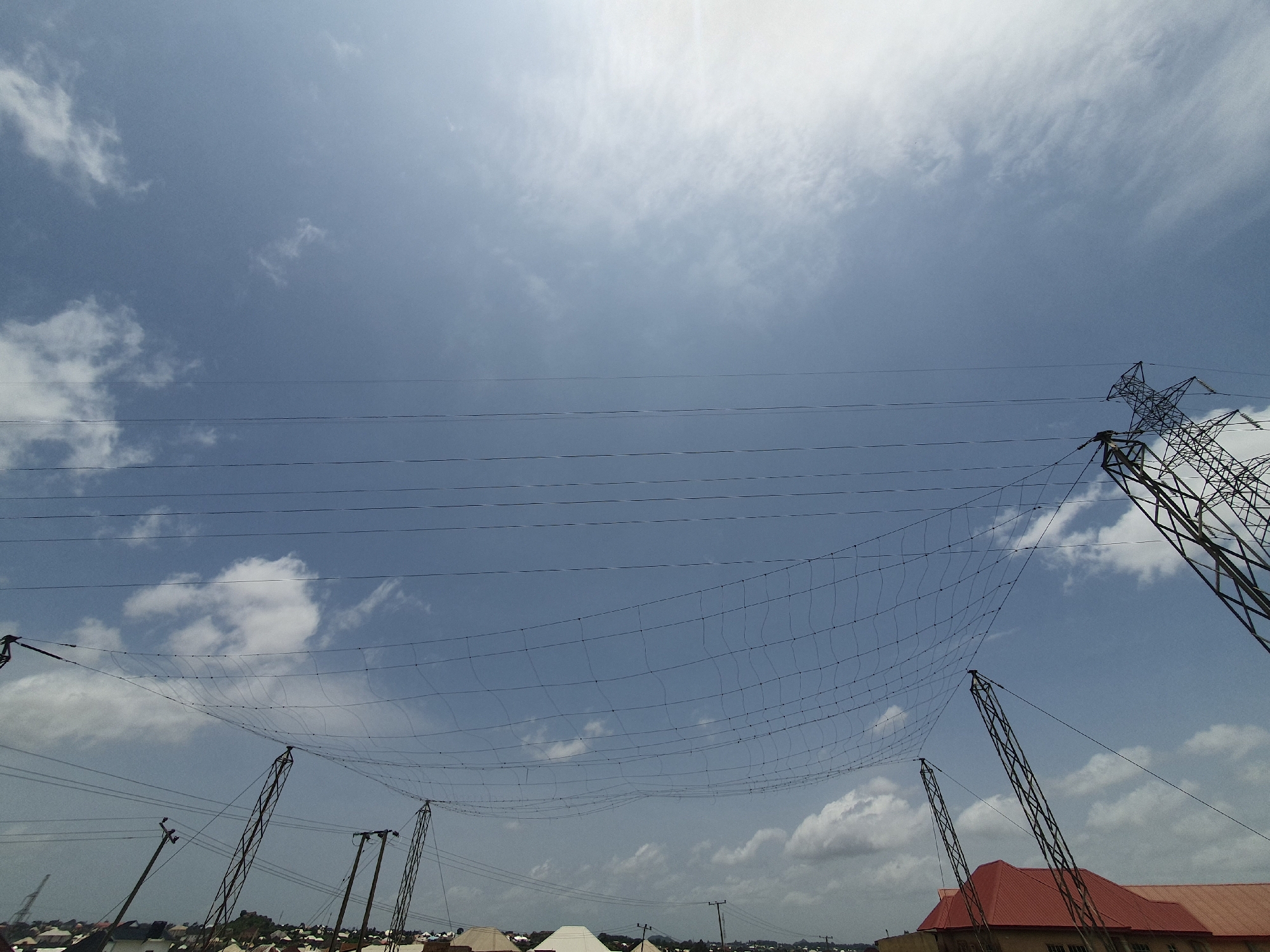 This is a wide-angle, low-angle shot of the sky above Gura Top, Nigeria. The sky is a hazy blue with scattered white clouds. Several power lines stretch across the frame, supported by metal towers of varying designs. One large, lattice-style tower is visible on the right, while smaller, more utilitarian poles are scattered throughout the lower portion of the image. Some of these poles appear to have multiple lines attached. A mesh-like netting is suspended between some of the poles, creating a rippling pattern against the sky. In the lower part of the image, the rooftops of buildings are visible, mostly red-tiled and some white-domed structures. The scene suggests an urban or semi-urban environment with significant electrical infrastructure.