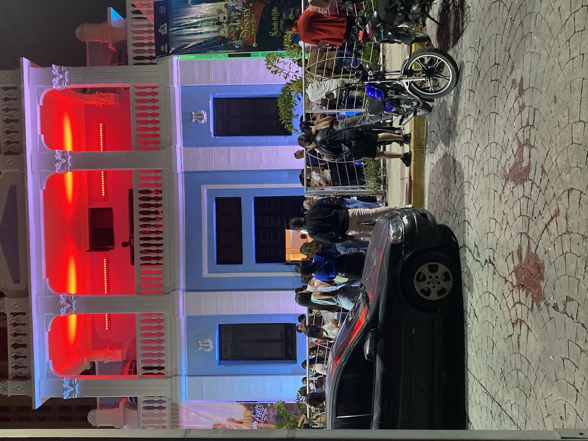 The image shows a street scene at night in Maracay, Venezuela, in front of a two-story building. The upper level of the building is illuminated with bright red lights, creating a vibrant and festive atmosphere. The lower level is painted light blue and features dark shutters on the windows. A crowd of people is gathered outside the building, many standing along a metal fence. In the foreground, a black car is parked, and a motorcycle is visible to the right. The ground is paved with cobblestones. There are some red stains on the cobblestones in the lower left portion of the image. A large banner with Disney characters is hanging on the right side of the building, advertising "Disney Magic Fest" for Saturday, July 11th. The lighting suggests it is evening or night.