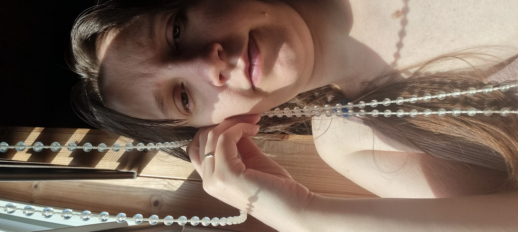 The image features a woman in a close-up, artistic shot, appearing to be lying on her side, looking directly at the viewer. Her head is resting gently on her right hand, which wears a simple ring on one finger. She has shoulder-length dark hair, a clear complexion, and her expression is soft and relaxed, with a slight, gentle smile. Her lips are a natural pink hue.
Strong, natural sunlight illuminates her face and upper body, creating pronounced shadows across her features and casting distinct, parallel striped shadows onto the wooden surface below her. These shadows suggest she is indoors, near a window with blinds or louvers, during a bright, sunny day, likely morning or late afternoon given the warm quality of the light.
A long strand of clear, spherical beads, possibly a decorative garland or a multi-strand necklace, is draped across her, with a portion running over her hair and shoulder, and another section held loosely by her left hand which is out of frame. A small blue bead is visible among the clear beads near her neck. The background shows a dark, indistinct area to the upper left and a light-colored wooden surface to the bottom and right, with more of the patterned shadows. The image is presented from a tilted perspective, rotated approximately 90 degrees clockwise. There is no visible text in the image. The setting is likely an indoor environment in Dunnae-myeon, South Korea.