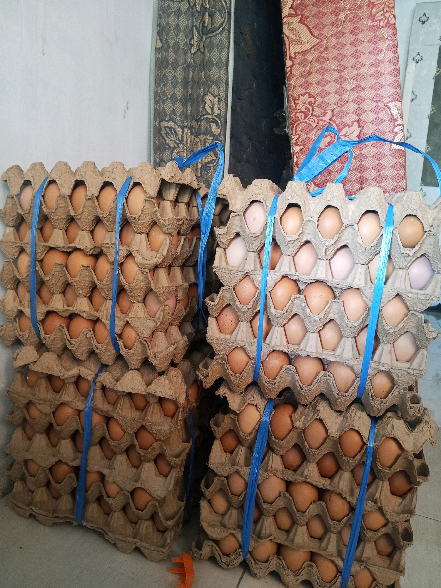 A stack of brown egg cartons, bound together with blue plastic straps, sits on a light-colored floor. Each carton is filled with brown and white eggs. In the background, patterned fabric hangs on what appears to be a wall. The overall setting suggests a storage area or a marketplace in Jinang Kampung Pansur, Indonesia.