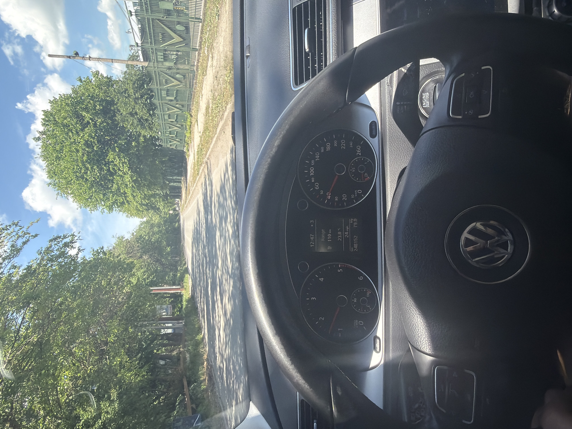 The image captures a driver's perspective from inside a Volkswagen vehicle, looking out onto a road bordered by trees and a distinctive green fence. The photo appears to have been taken in portrait mode, with the dashboard and steering wheel on the right, and the external view on the left.
Inside the car, the black steering wheel, featuring the Volkswagen logo, is prominent. Behind it, the instrument cluster is visible, displaying a speedometer (reading up to 260 km/h, currently at 0) and a tachometer (up to 6, also at 0). A digital screen within the cluster shows the time as "12:47", indicating midday. Other readings include "Range 110 km", an external temperature of "23.0 °C", a "trip" distance of "79.0", and an odometer reading of "248152". A fuel gauge shows the tank is approximately half full. An "ENGINE START/STOP" button is visible near the steering column.
The exterior view shows a clear, sunny day with a blue sky dotted with white clouds. A paved road is visible, with strong shadows cast upon it, consistent with midday. Lush green trees line both sides of the road. On the left side of the frame, a tall, elaborate green metal fence or gate with geometric patterns stands prominently, suggesting a property boundary or public works structure. A utility pole with equipment is also visible near the fence. The overall scene suggests a pleasant, calm environment, likely in a rural or suburban area of Shumen, Bulgaria.
