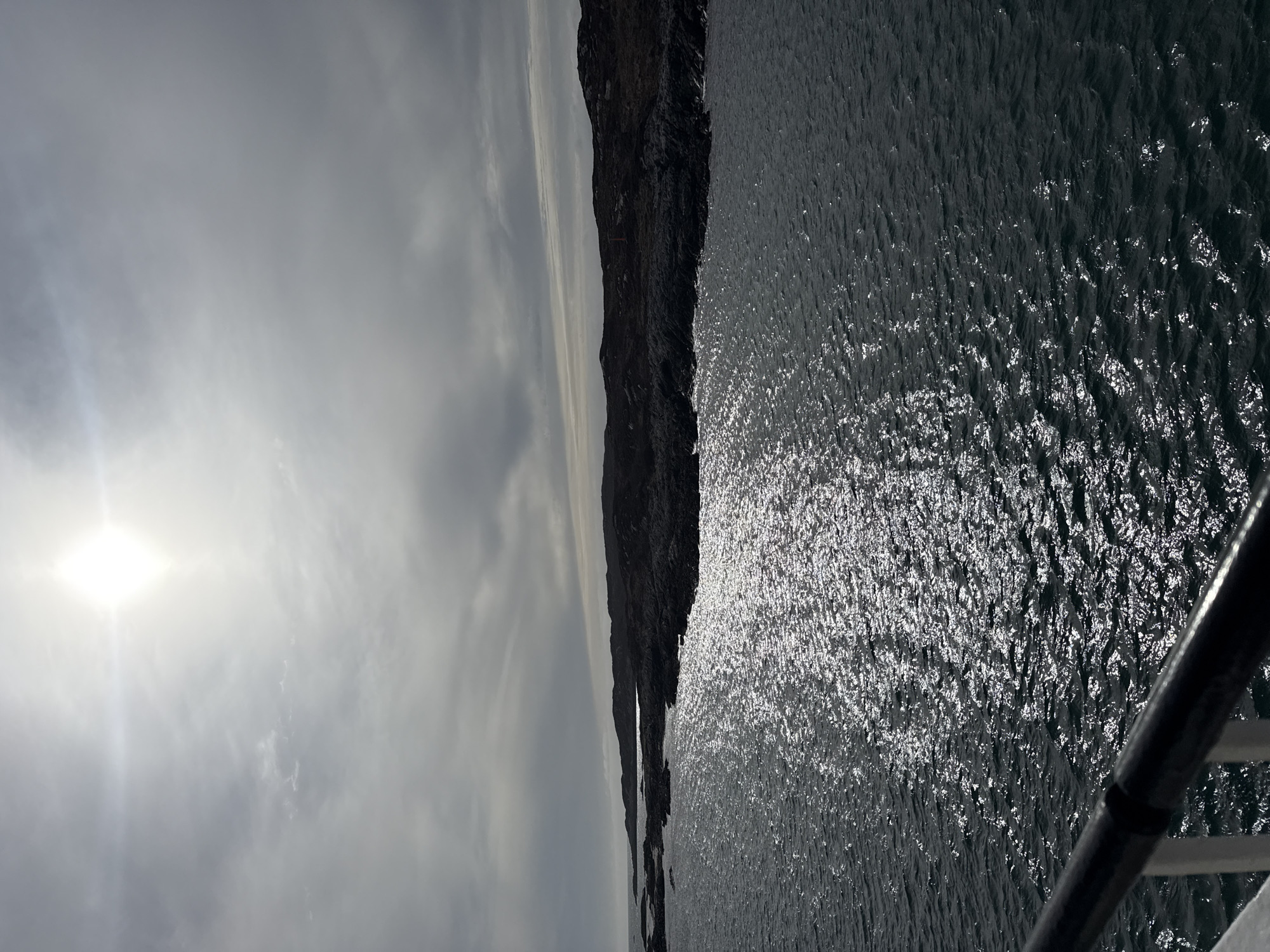 The image depicts a body of water glistening under sunlight near a rocky cliff. Overcast skies dominate the top left of the frame. A portion of a metal railing is visible in the bottom right. The location appears to be coastal, but the specific city and region are unknown.